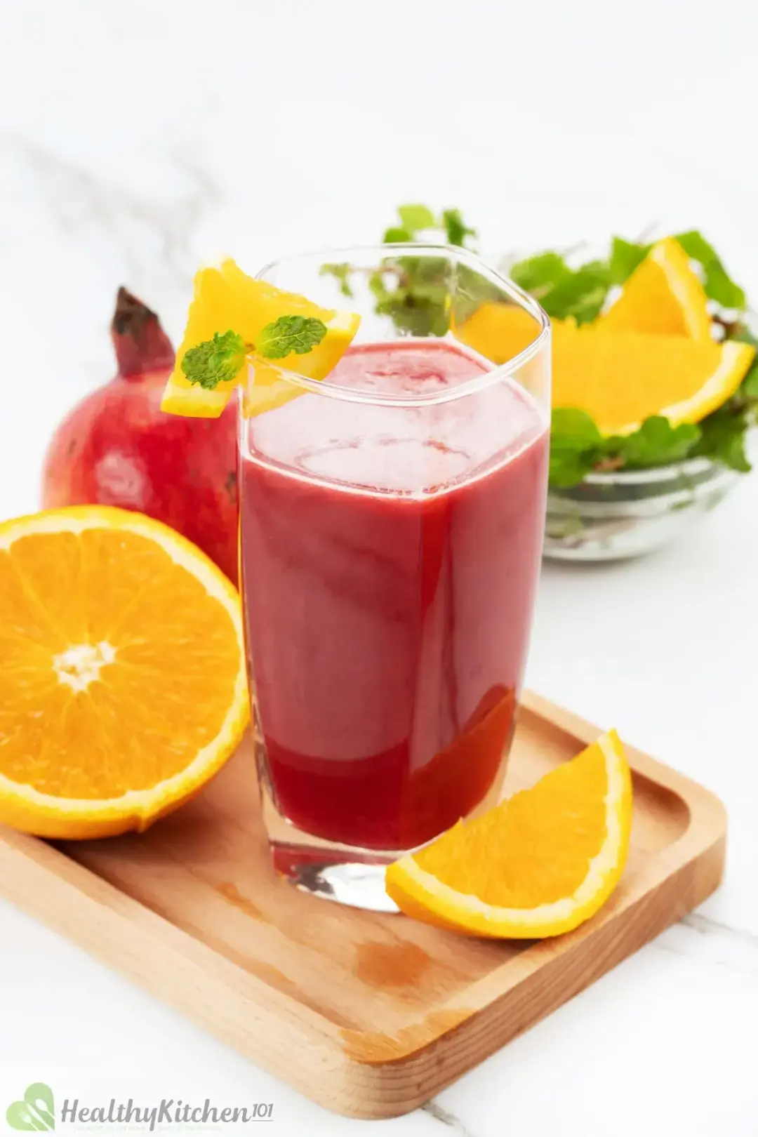 A glass of pomegranate orange juice put on a wooden board with orange slices, with some more fruits in the background