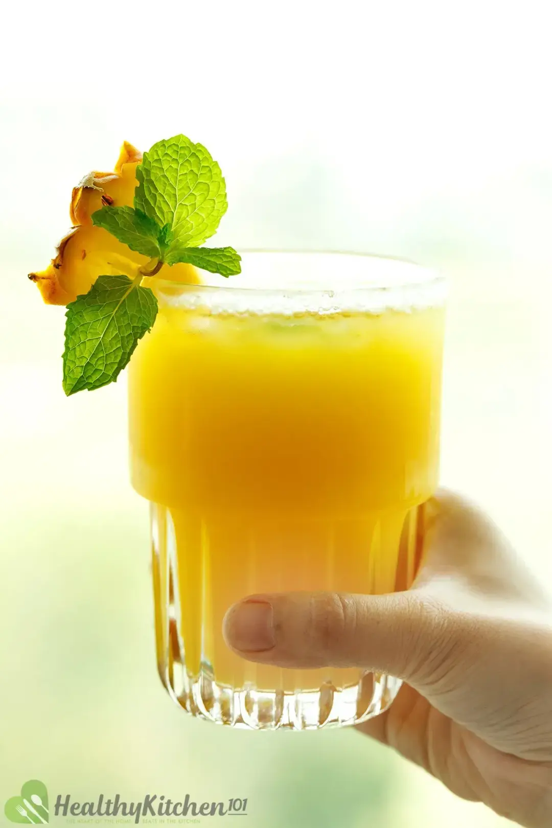 A hand holding a glass of pineapple juice and cider cocktail near a window