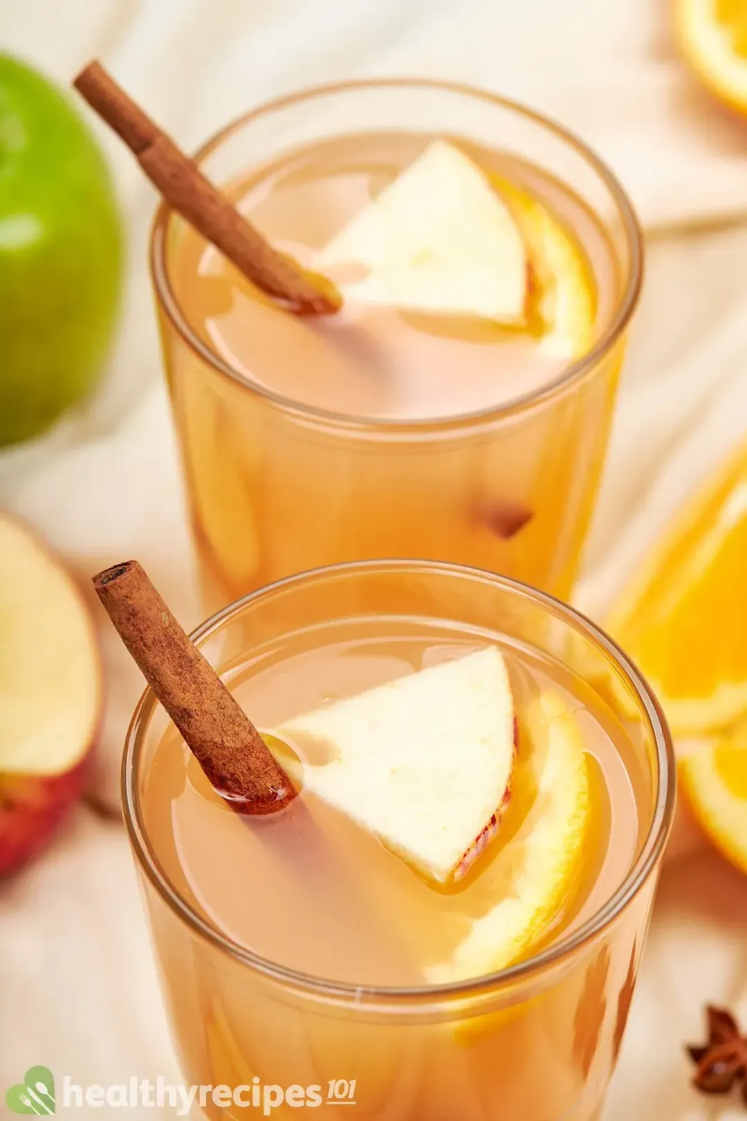 Two glasses of apple cider, topped with apple triangles and cinnamon sticks