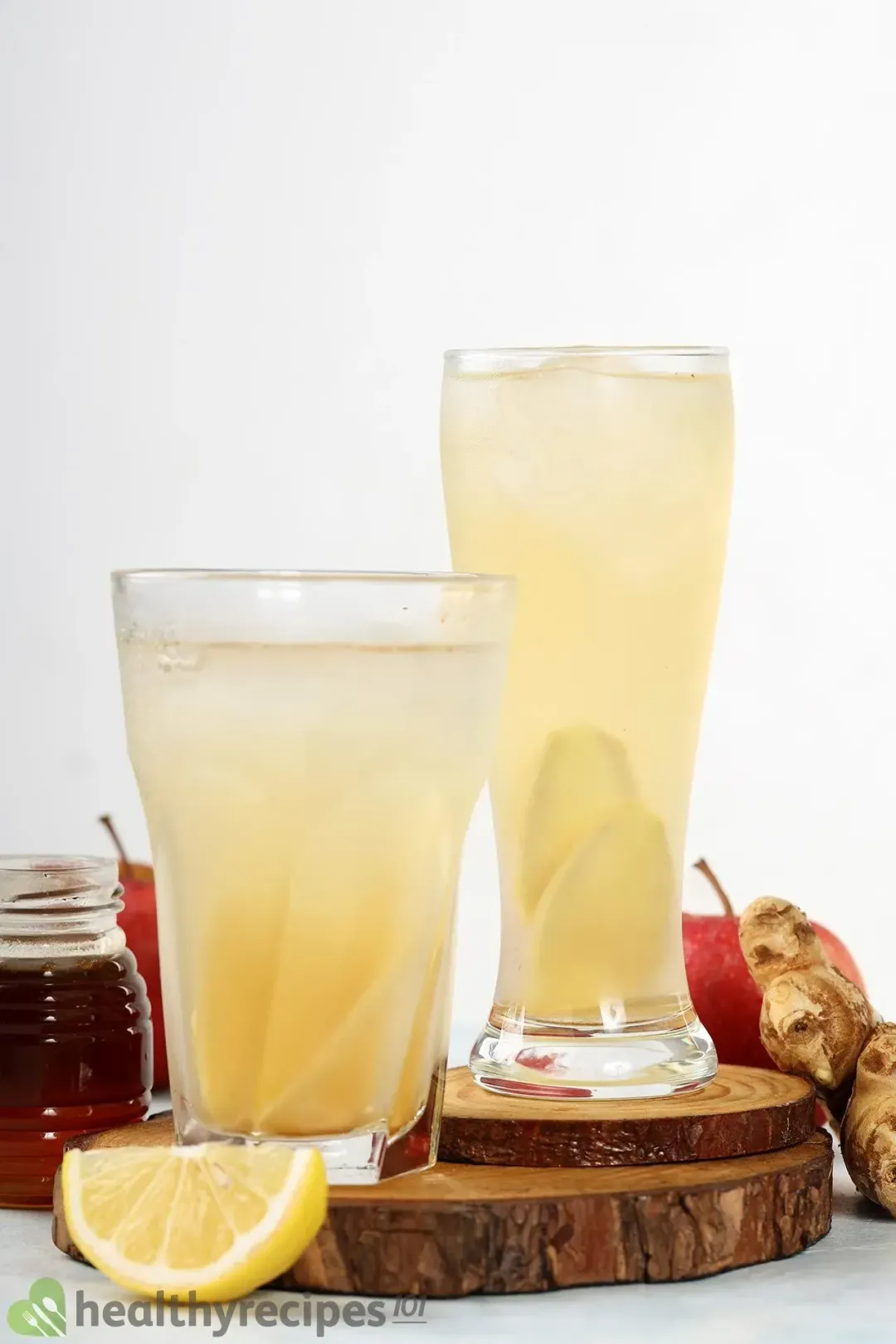 Two tall glasses of apple cider vinegar drinks with ginger slices inside, next to citruses and a jar of honey
