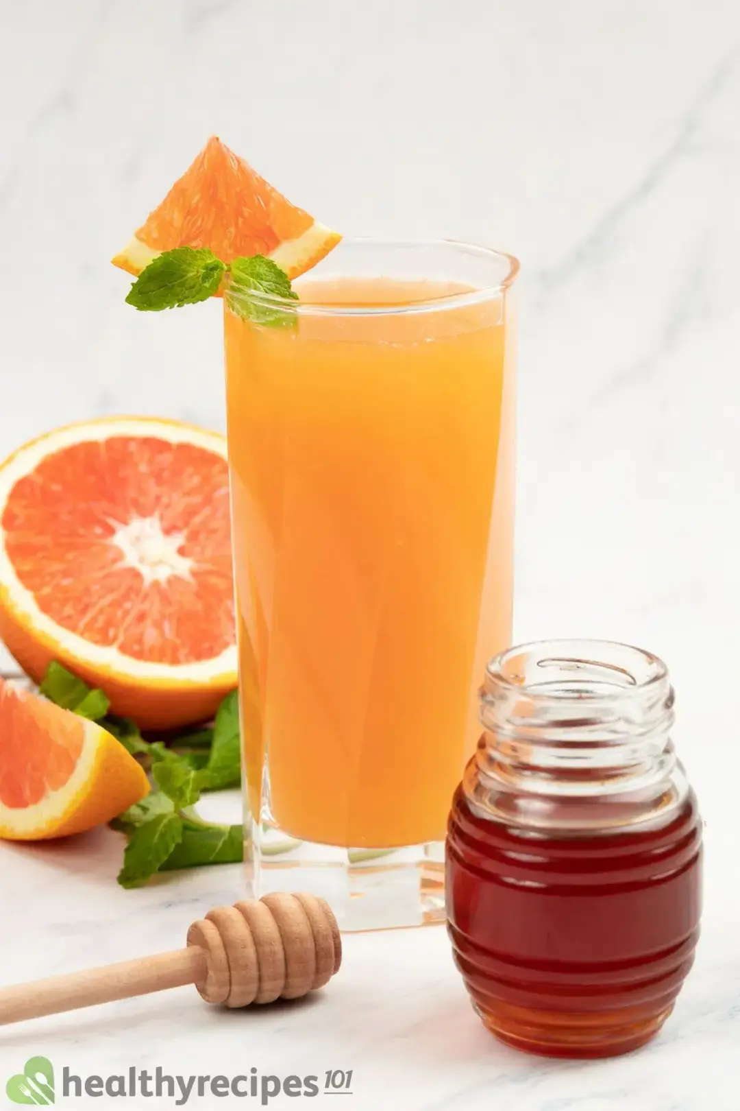 A glass of grapefruit juice garnished with mints, next to half of a grapefruit and a honey jar