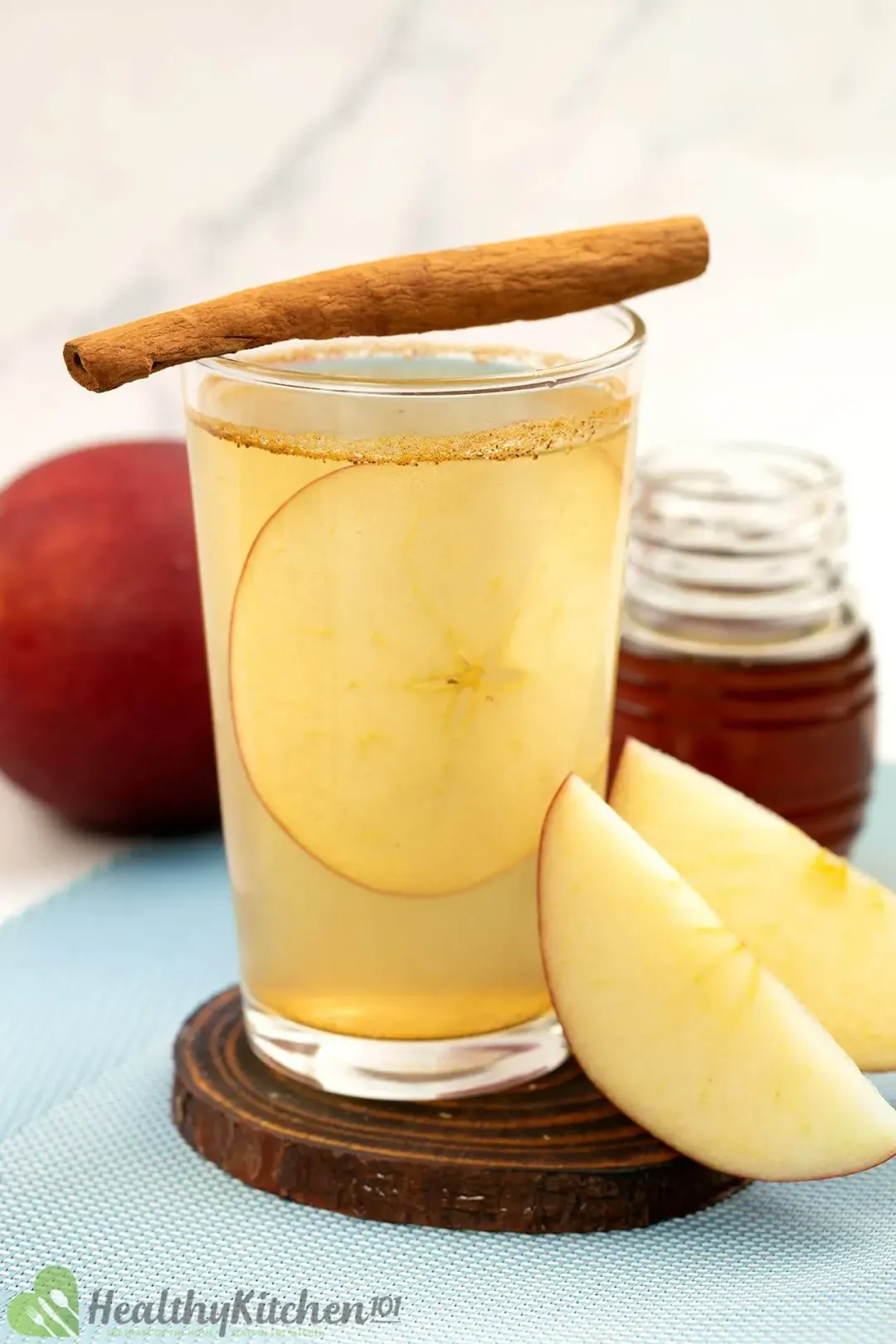 Next to apple wedges and below a cinnamon stick, a shot glass of apple honey cider with apple slice inside