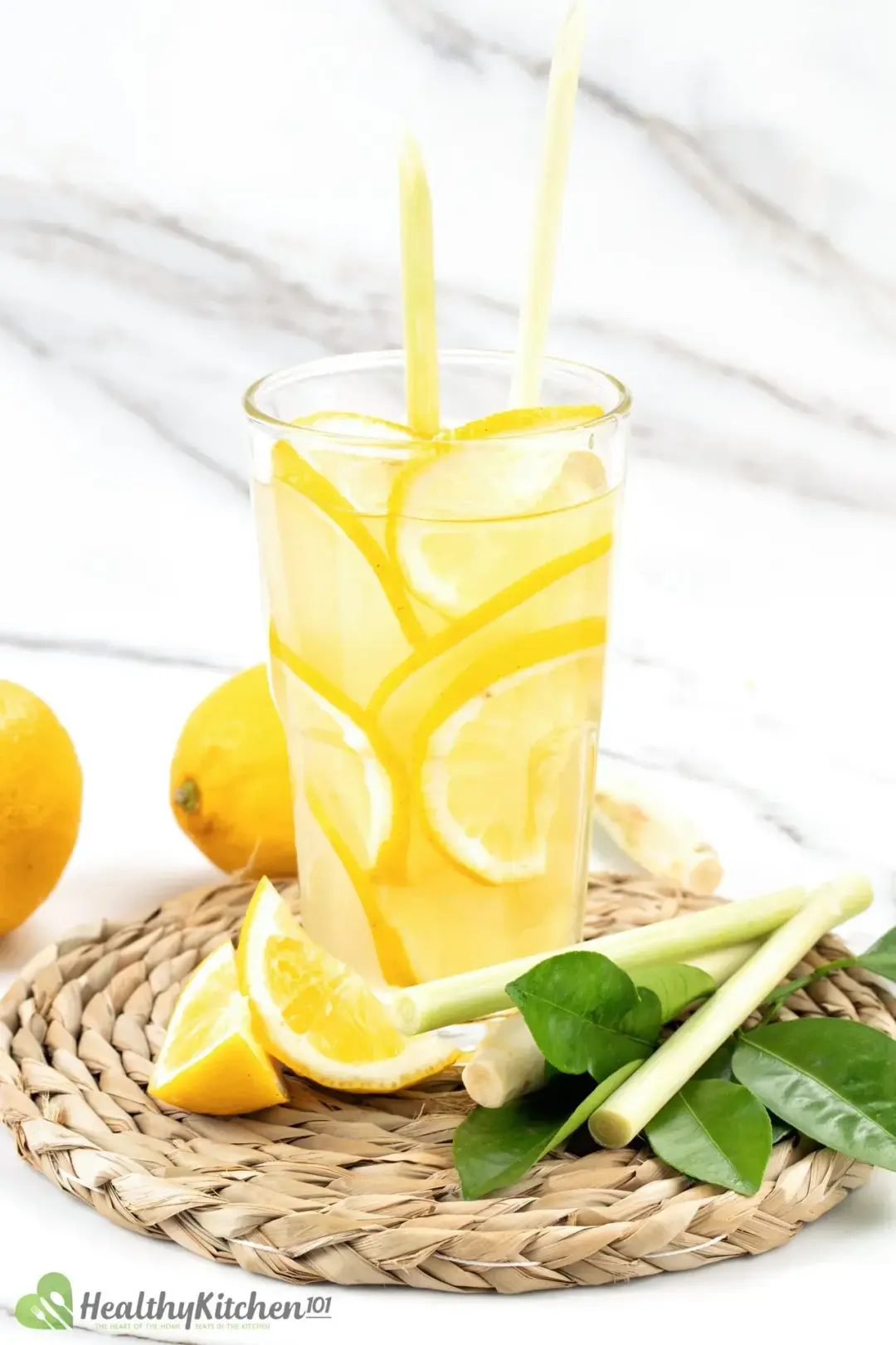 A glass full of juice and lemon wheels put on a woven coaster with lemon wedges, lemongrass stalks, and lemon leaves