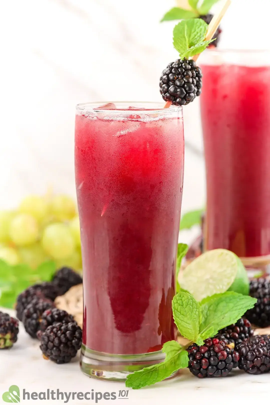 Two tall iced glasses of black berry juice drinks, garnished with mints and put next to grapes