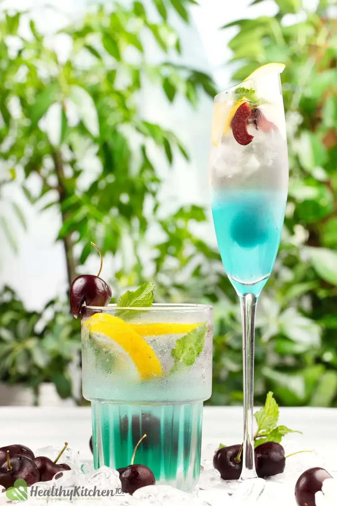 Two cocktail glasses of a cyan-hued drink, with cherries and lemon wedges all lying around