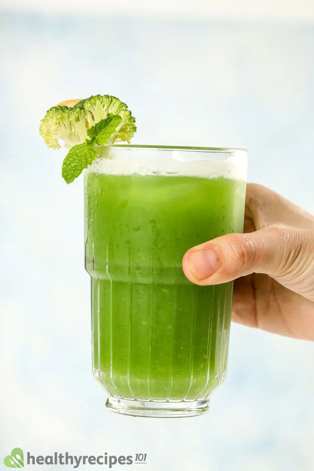 A hand holding a glass of broccoli juice drinks garnished with lime wedges and mints