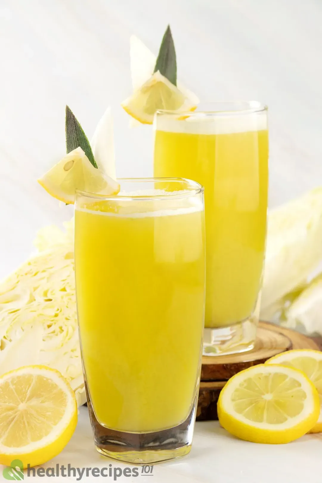 Two glasses of yellow-green cabbage drink, next to some cabbages and lemon wheels