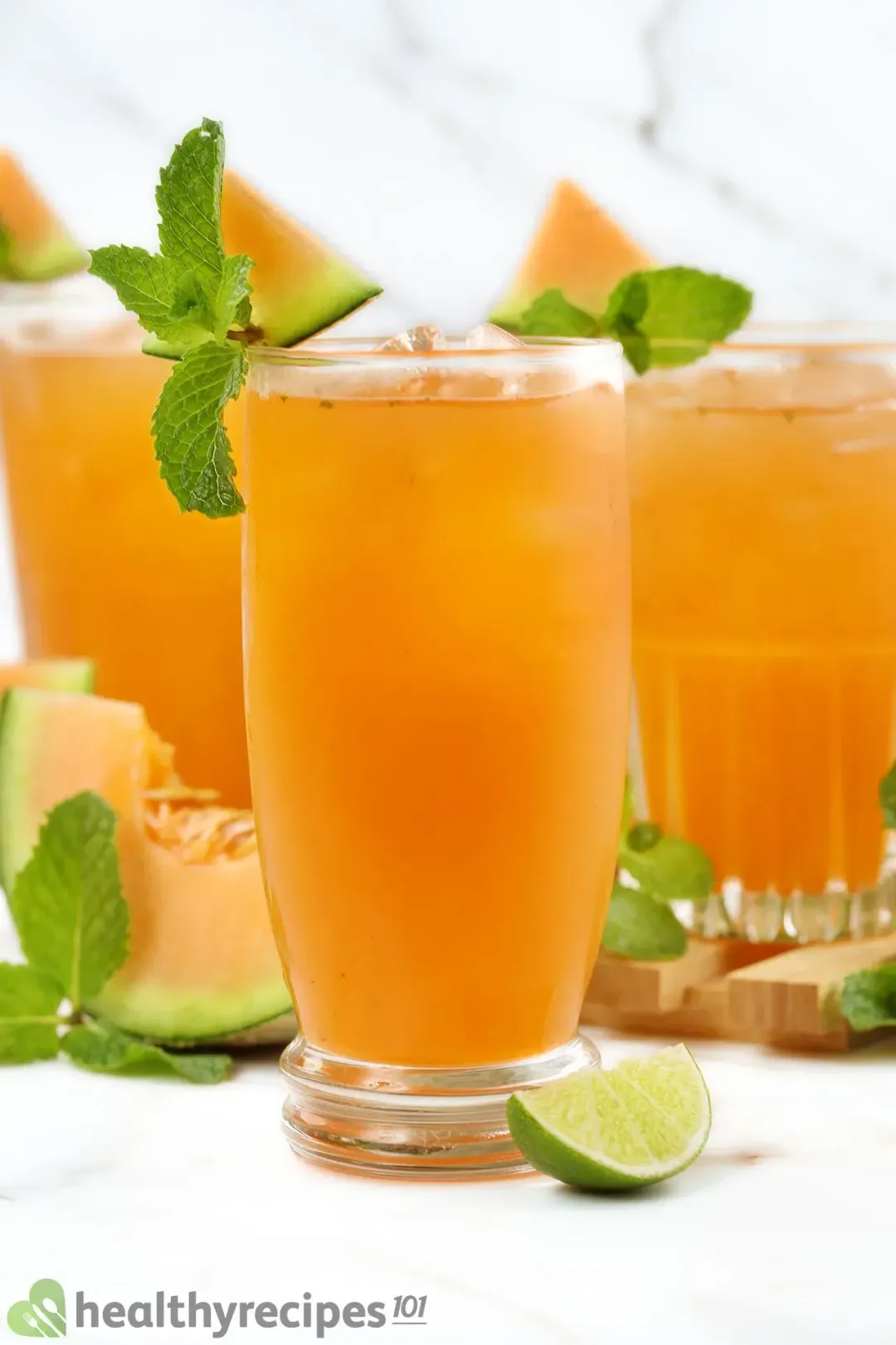 Three iced glasses of cantaloupe drinks, garnished with mints, cantaloupe wedges, and lime wedges lying around