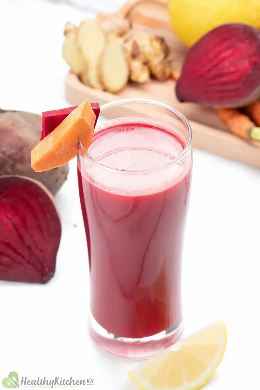 A tall glass of beetroot drink garnished with carrot slices, beetroot half, lemon wedges, and ginger knobs