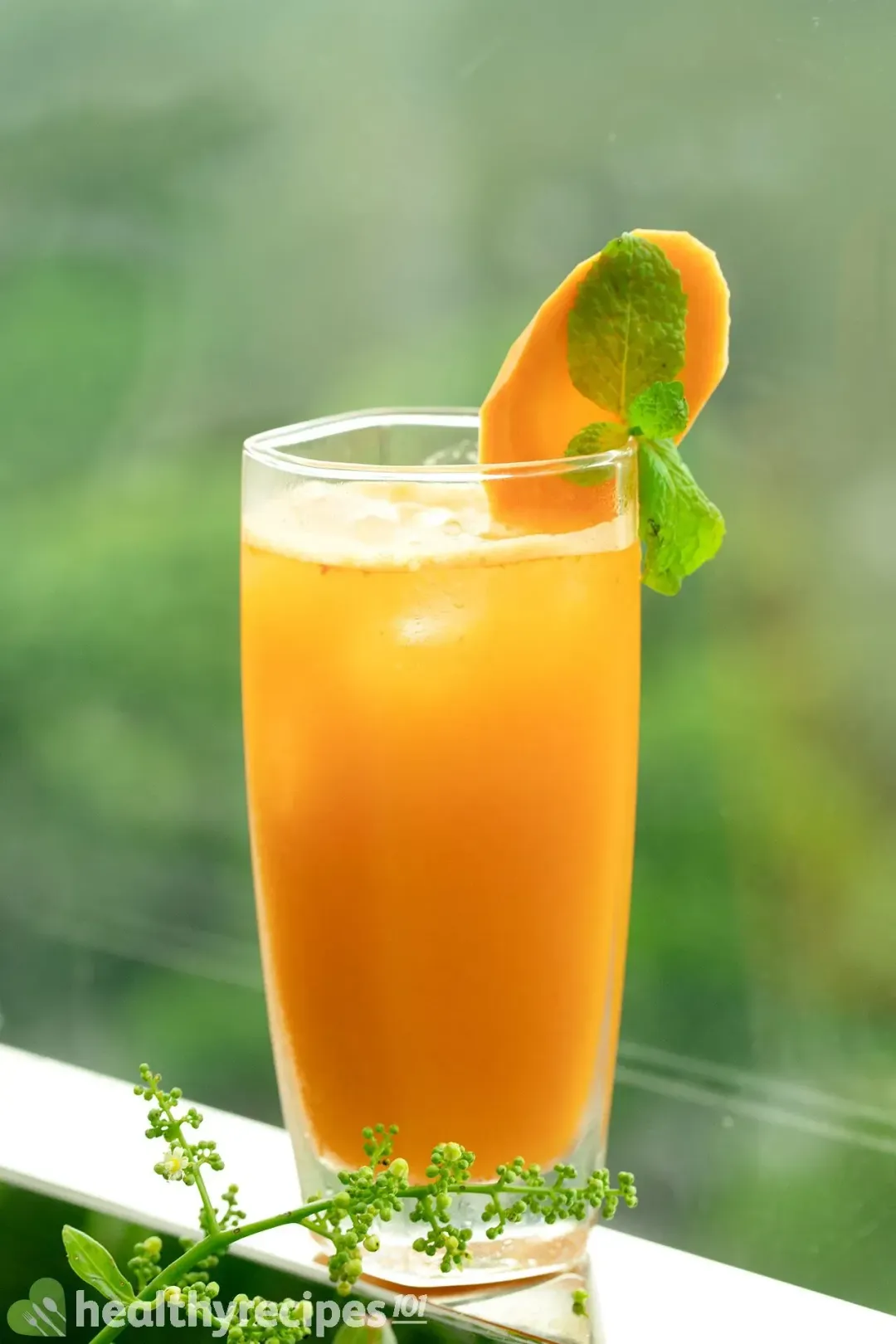 A tall glass of ice and orange carrot drink with mints and carrots slices cucumber lime juice