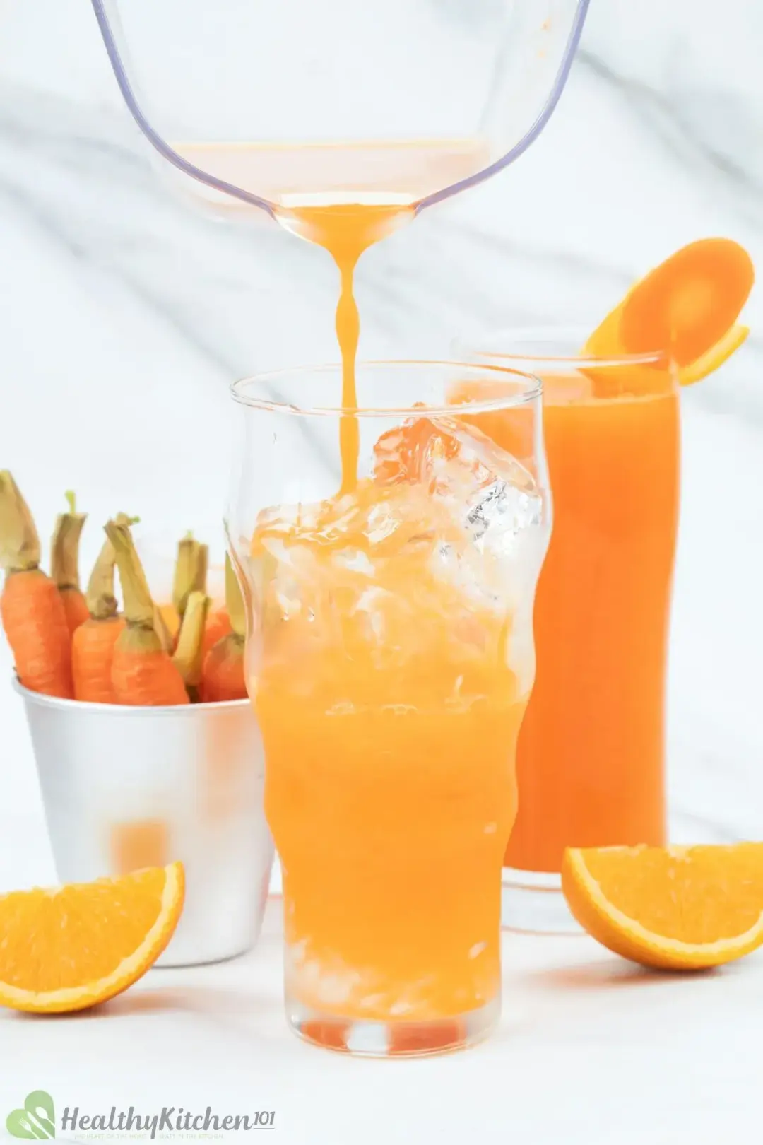 A glass pitcher pouring an orange drink into an iced glass, next to an orange glass, orange wedges, and baby carrots