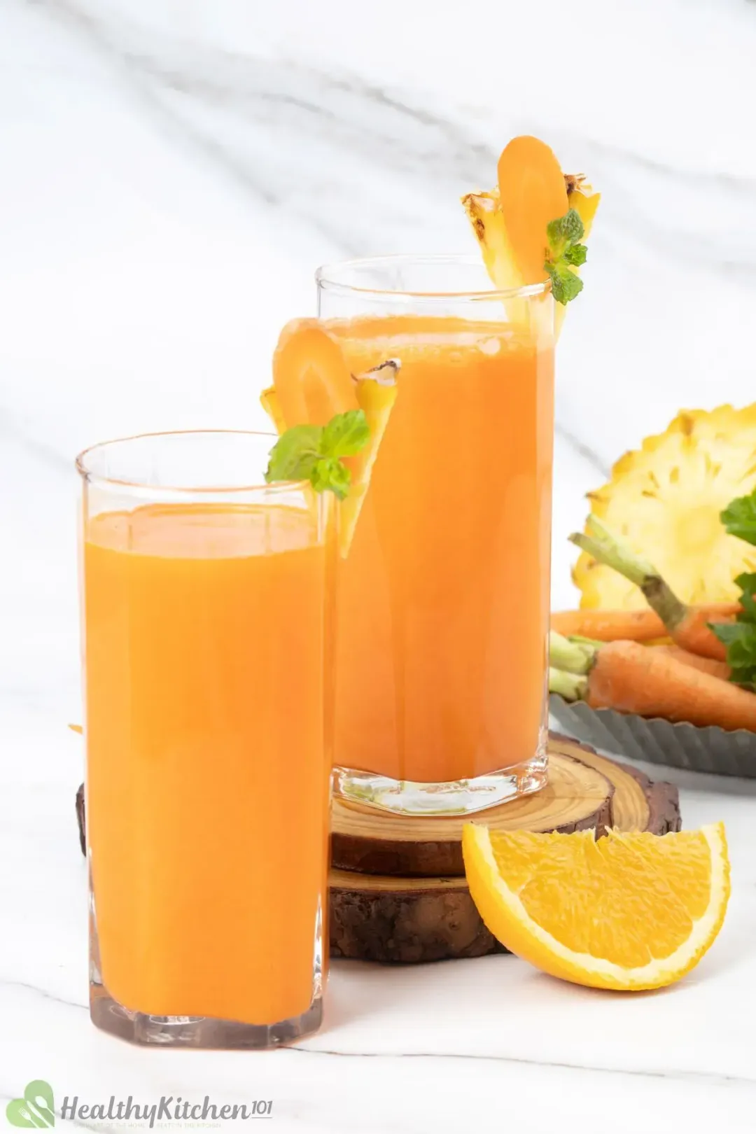Two glasses of pineapple orange carrot juice of different sizes garnished with carrot slices orange wedge and baby carrots
