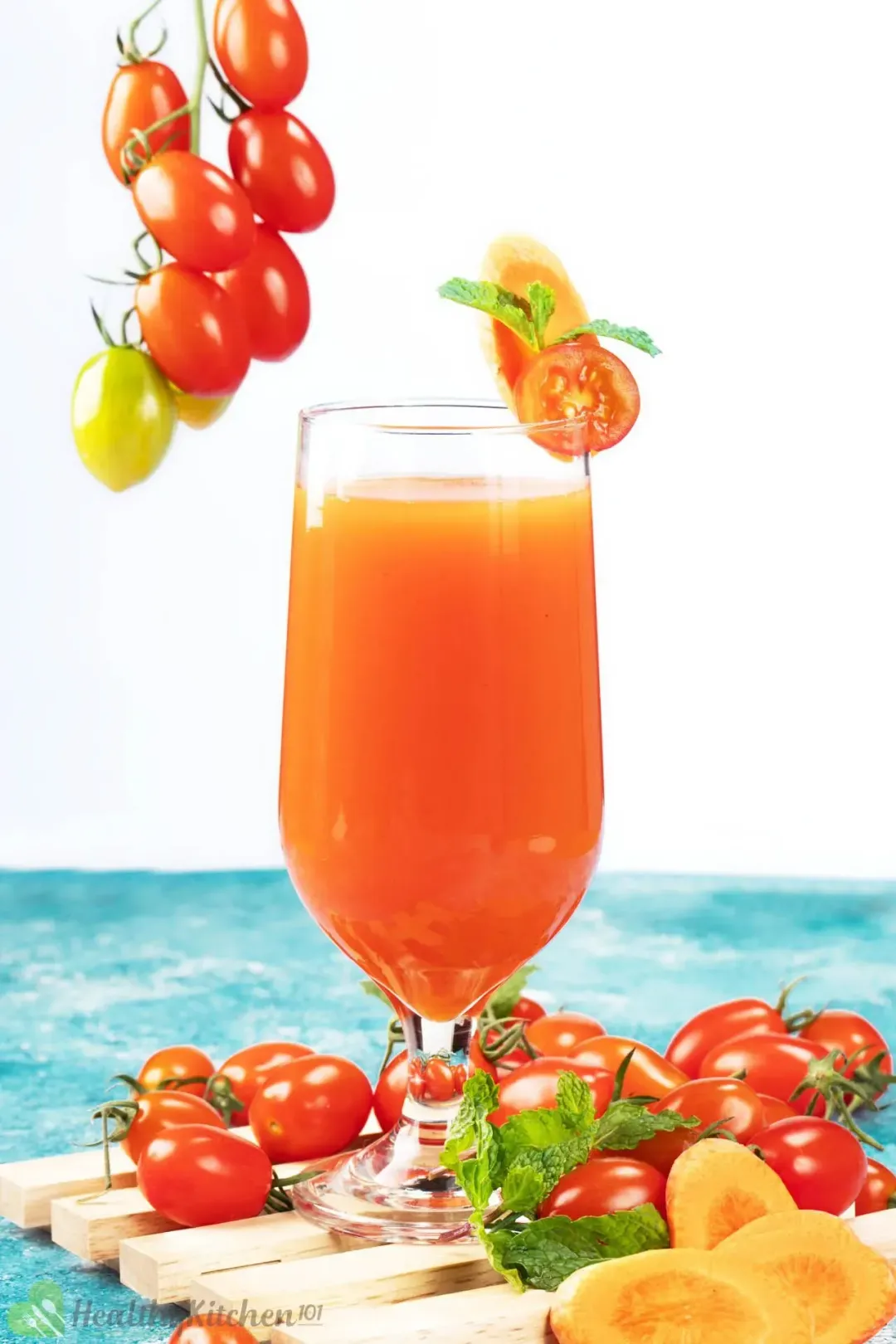 A glass of carrot tomato juice surrounded by lots of cherry tomatoes on vine, on a blue table