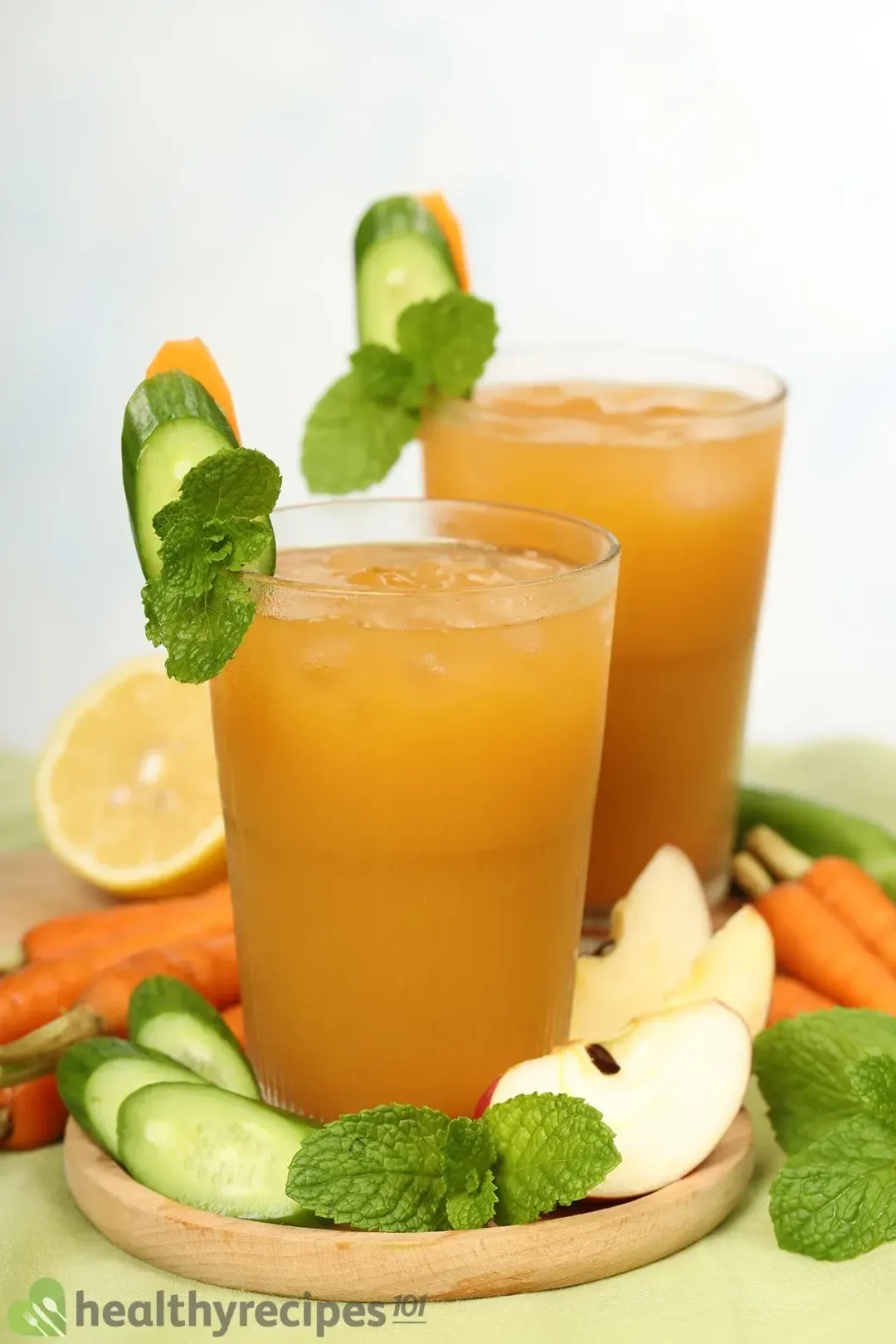 Two glasses of iced brown drink garnished with sliced cucumber, mint leaves, apple wedges, and baby carrots