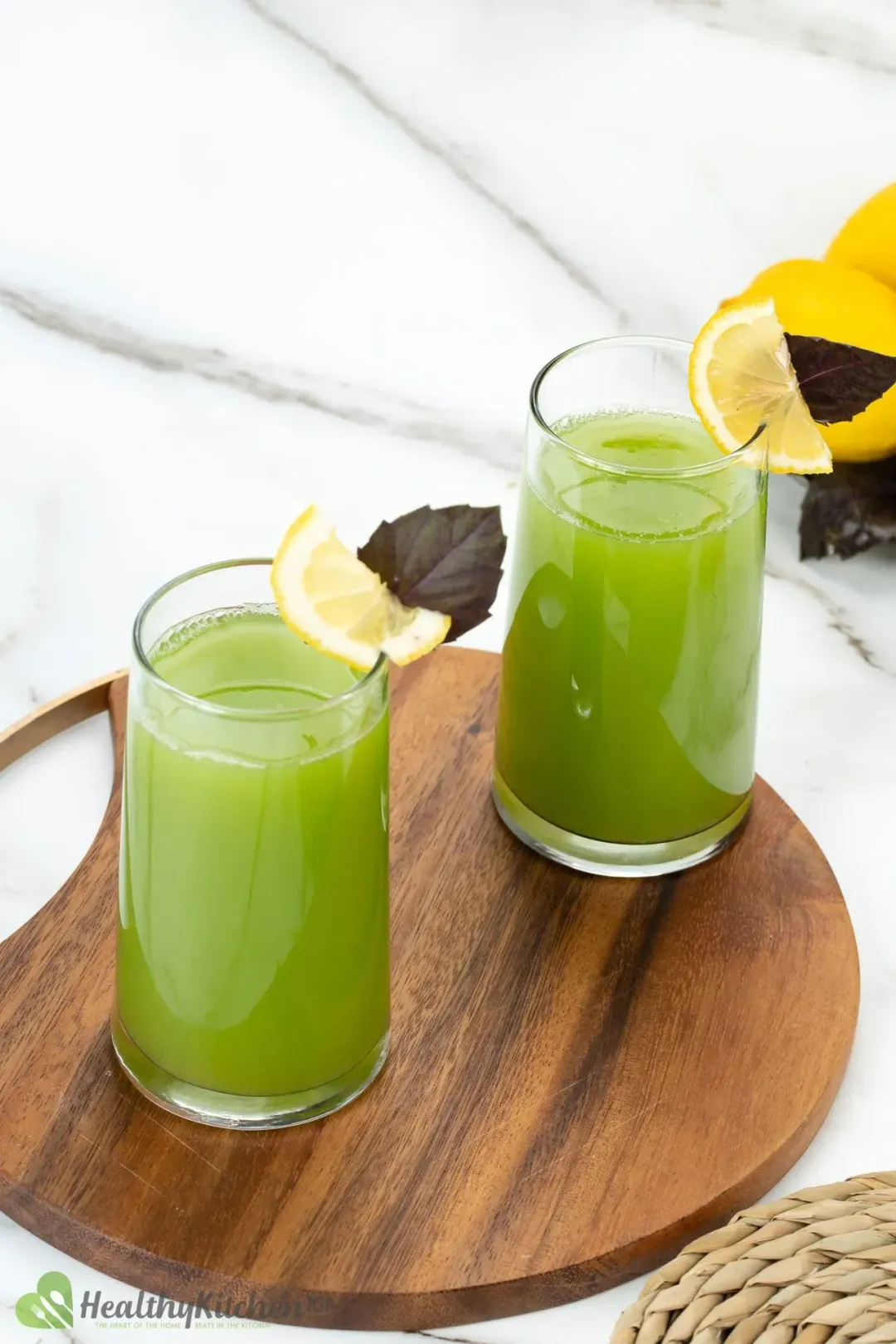 On a wooden plate, two glasses of cucumber lemon drinks both garnished with lemon wedges and purple leaves