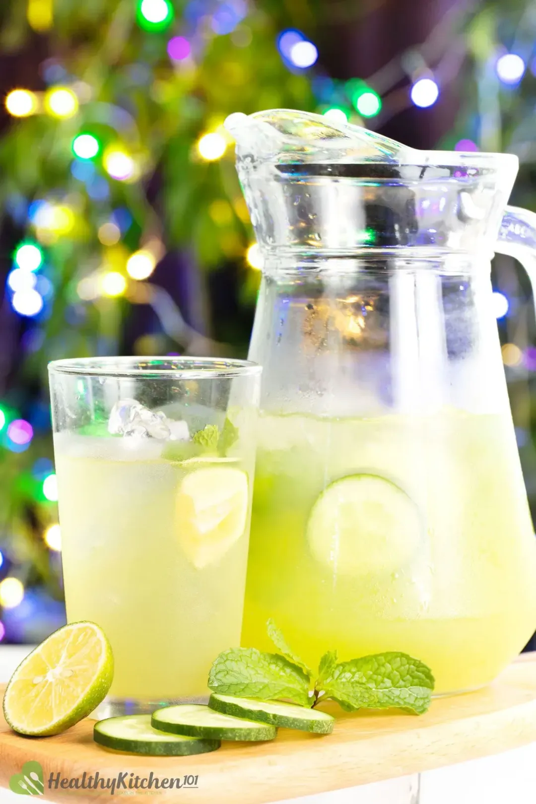 The pitcher of cucumber and lime juice next to a glass with ice and drink on a wooden board along with cucumber slices and a mint leaf