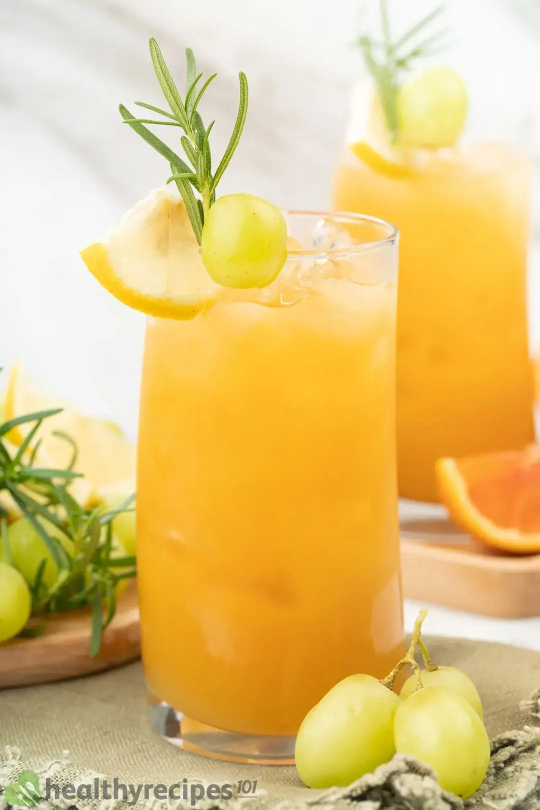 Glasses of grapefruit drink with ice, garnished with rosemary, grape, a lemon wedge, and whole grapes on the side