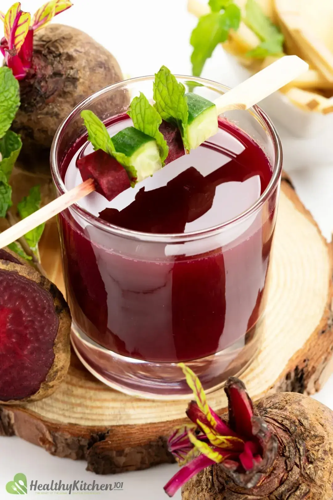 A short glass of beetroot juice topped with a skewer of cubed cucumber, beetroot, and mint leaves, next to whole beetroots