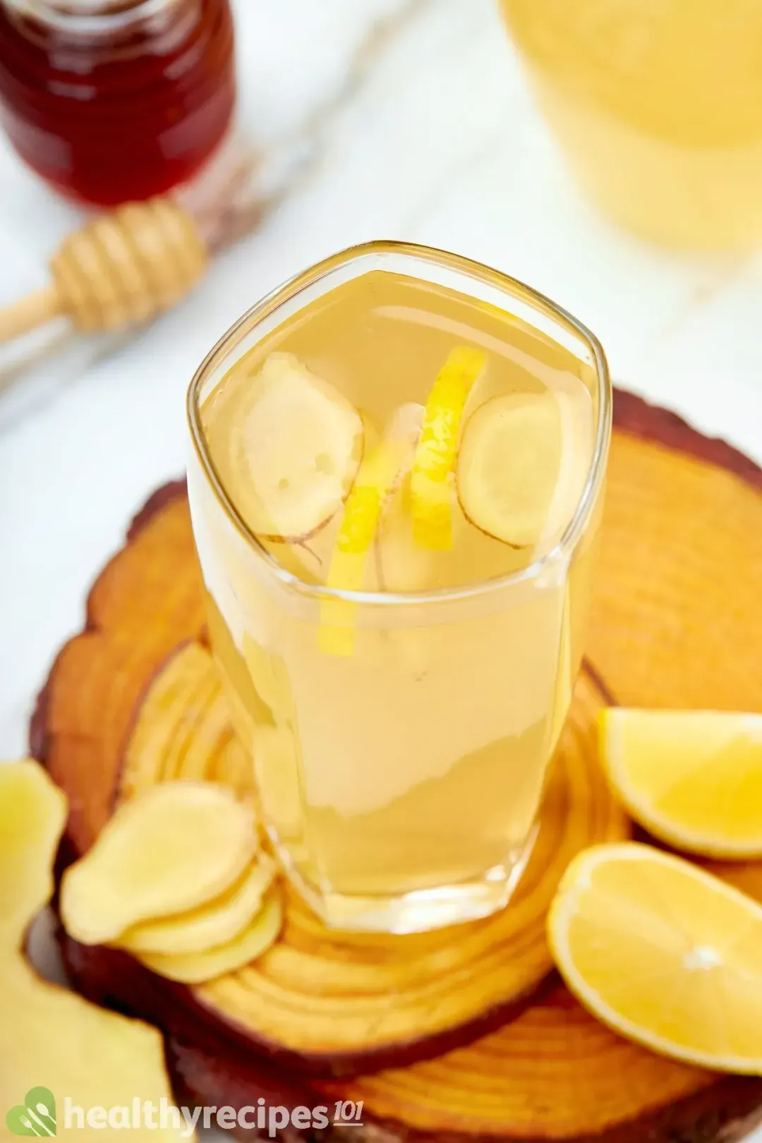 A tall glass of drink filled with sliced ginger, lemons, and next to some sliced ginger and wedged lemons