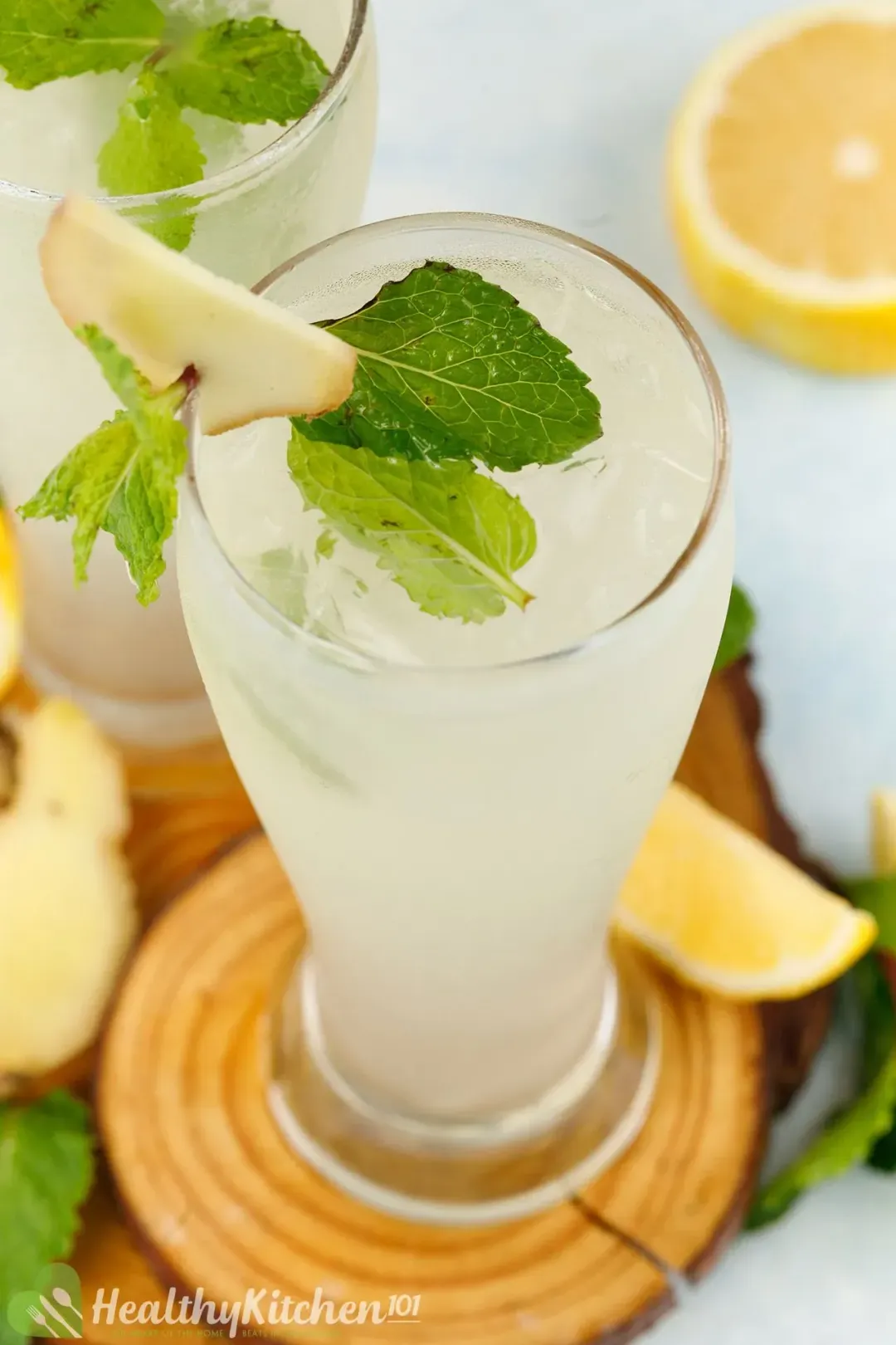 A tall glass of iced drink topped with mint leaves, ginger slice, and put next to some lemon wedges