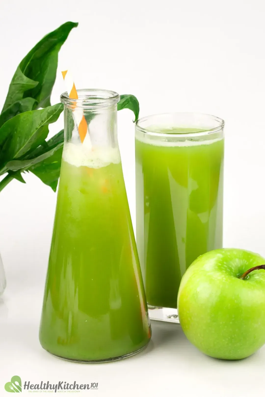  A pitcher and a glass of green apple juice decorated with an orange-striped straw alongside a green apple and spinach