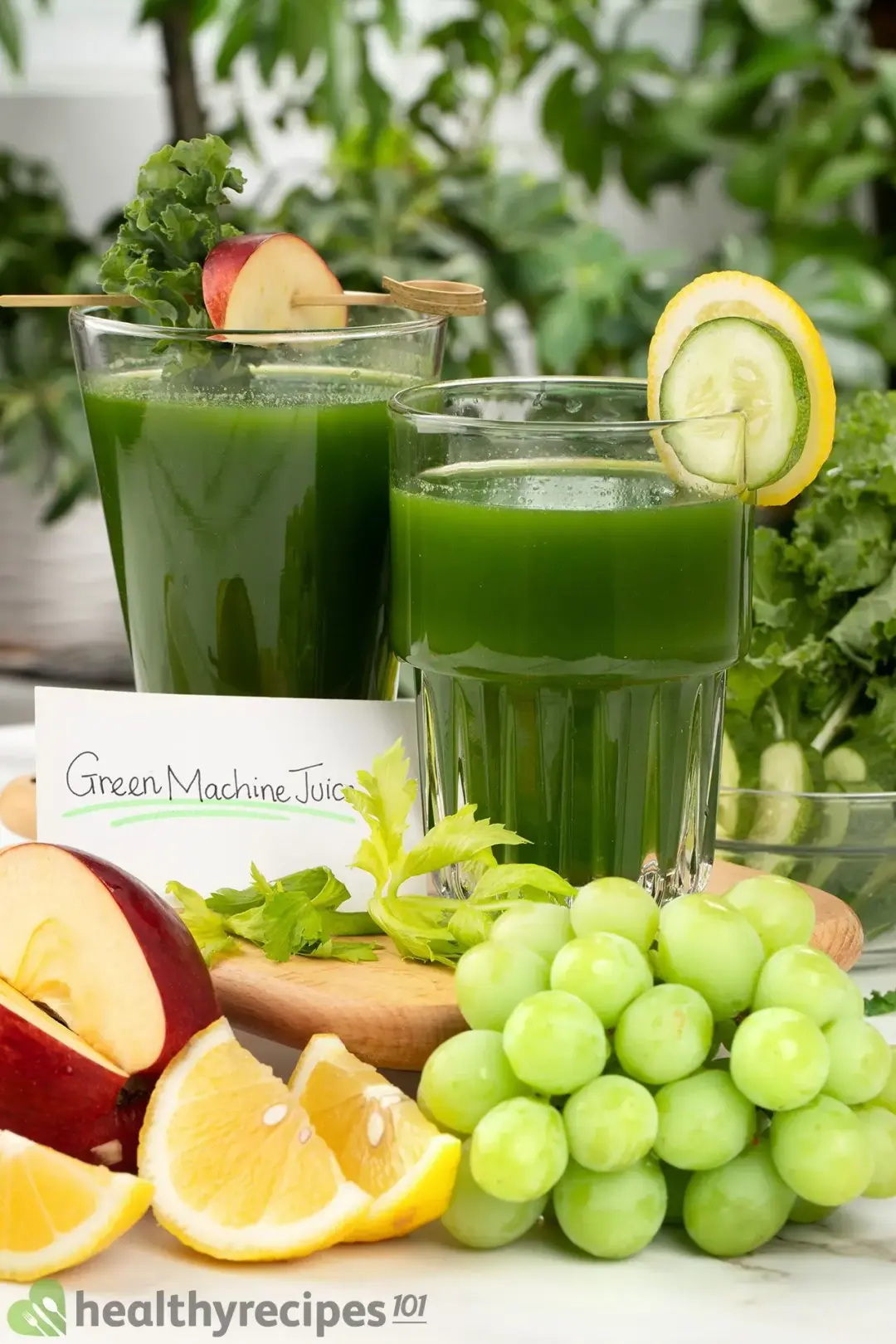 Two glasses of green machine juice with a note on the side placed on a wooden board, with one decorated with kale leaves and a slice of apple, and another decorated with a cucumber slice and a lemon slice