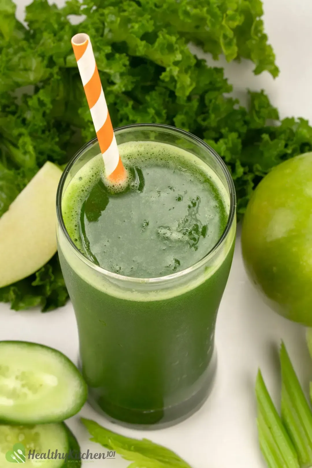A glass of green vegetable juice whose picture is taken from above, with an orange straw dunked in and green fruits and vegetables in the background