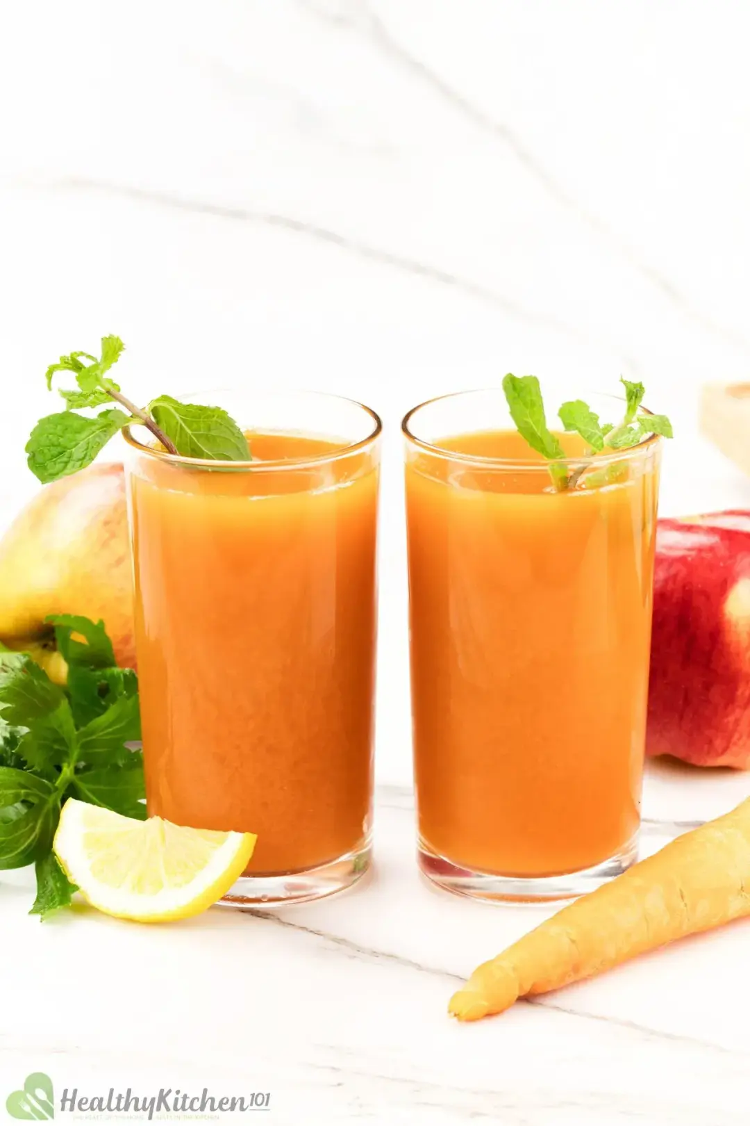 Two similar carrot juice glasses with a mint sprig in each and next to a carrot, apples, and lemon wedge
