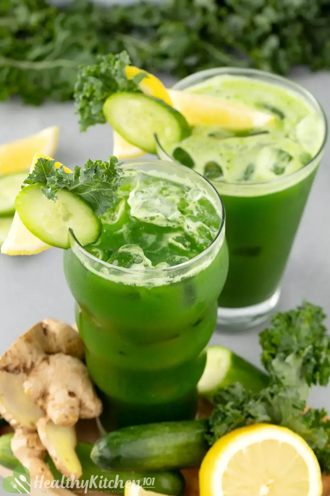 Two glasses of green kale juice with ice, garnished with kale leaves, cucumber slices, ginger, and lemons