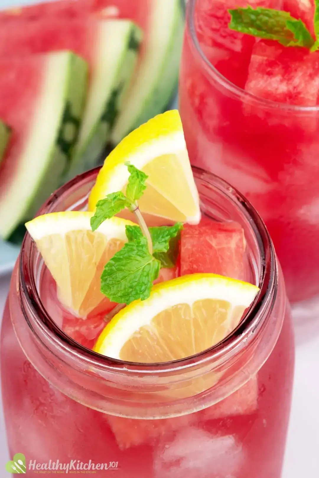 A close-up shot of a jar of watermelon juice and lemon garnished with three slices of lemon and mint leaves with a plate of watermelon slices and a glass of watermelon juice and lemon on the background.