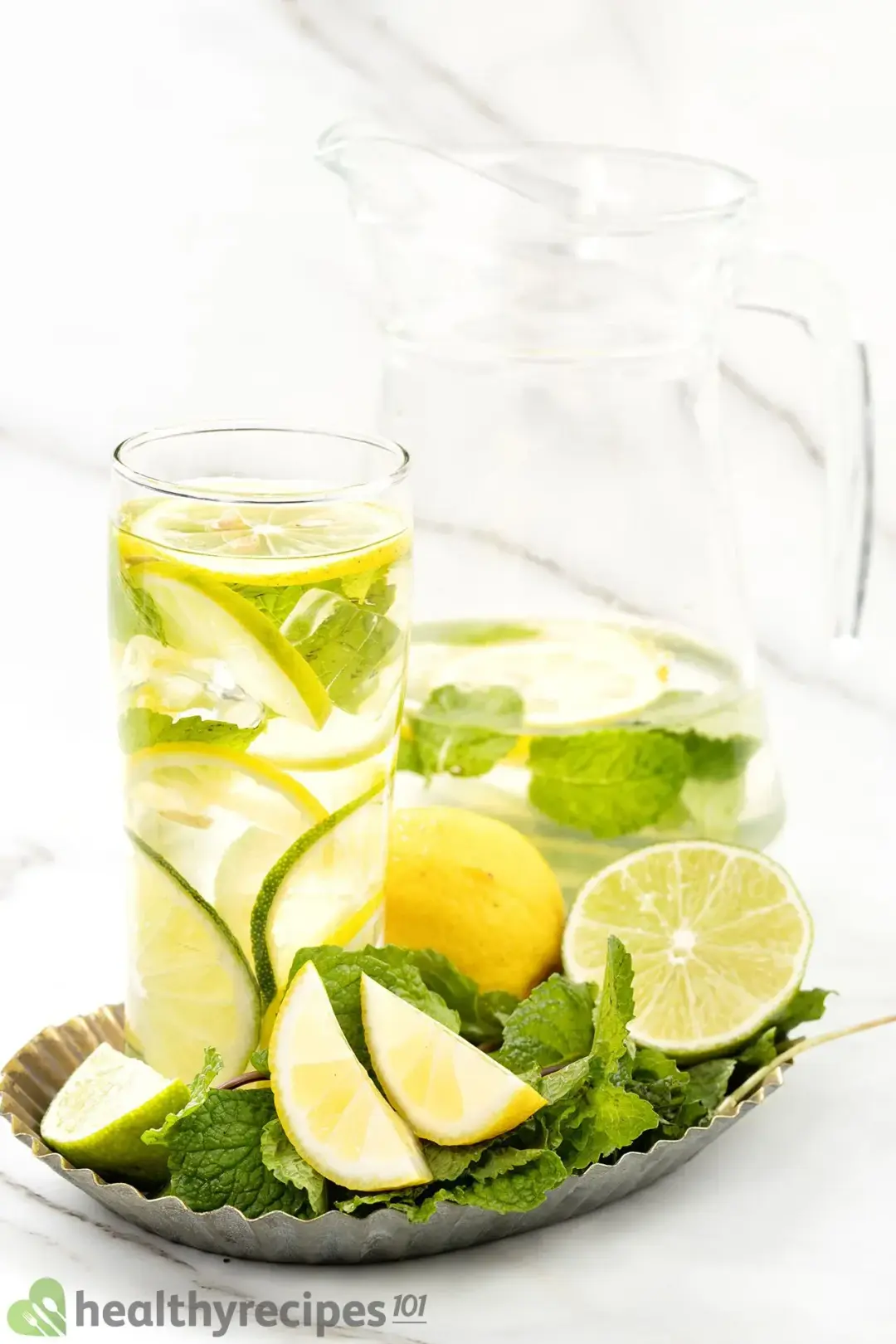 A glass and a pitcher of water full of citrus wheels, on a tray with lemon wedges and lots of mint leaves