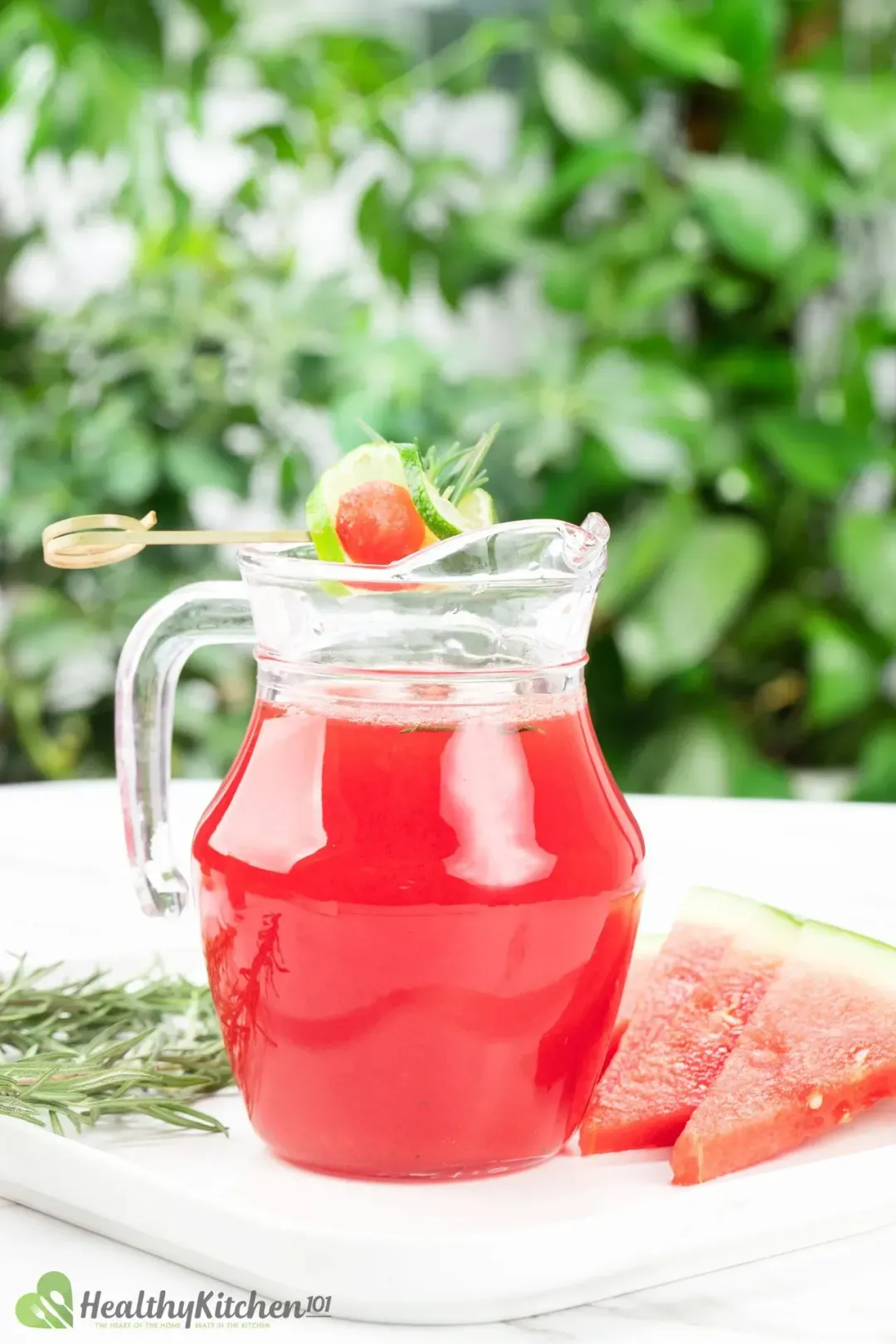 A watermelon juice pitcher, watermelon wedges, and rosemary sprigs on a white table out in the garden