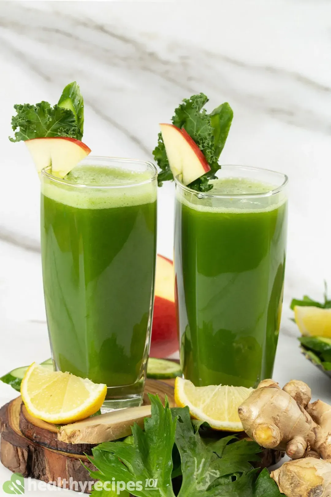 Two glasses of mean green juice put side by side, surrounded by lime wedges, green leaves, and ginger knobs