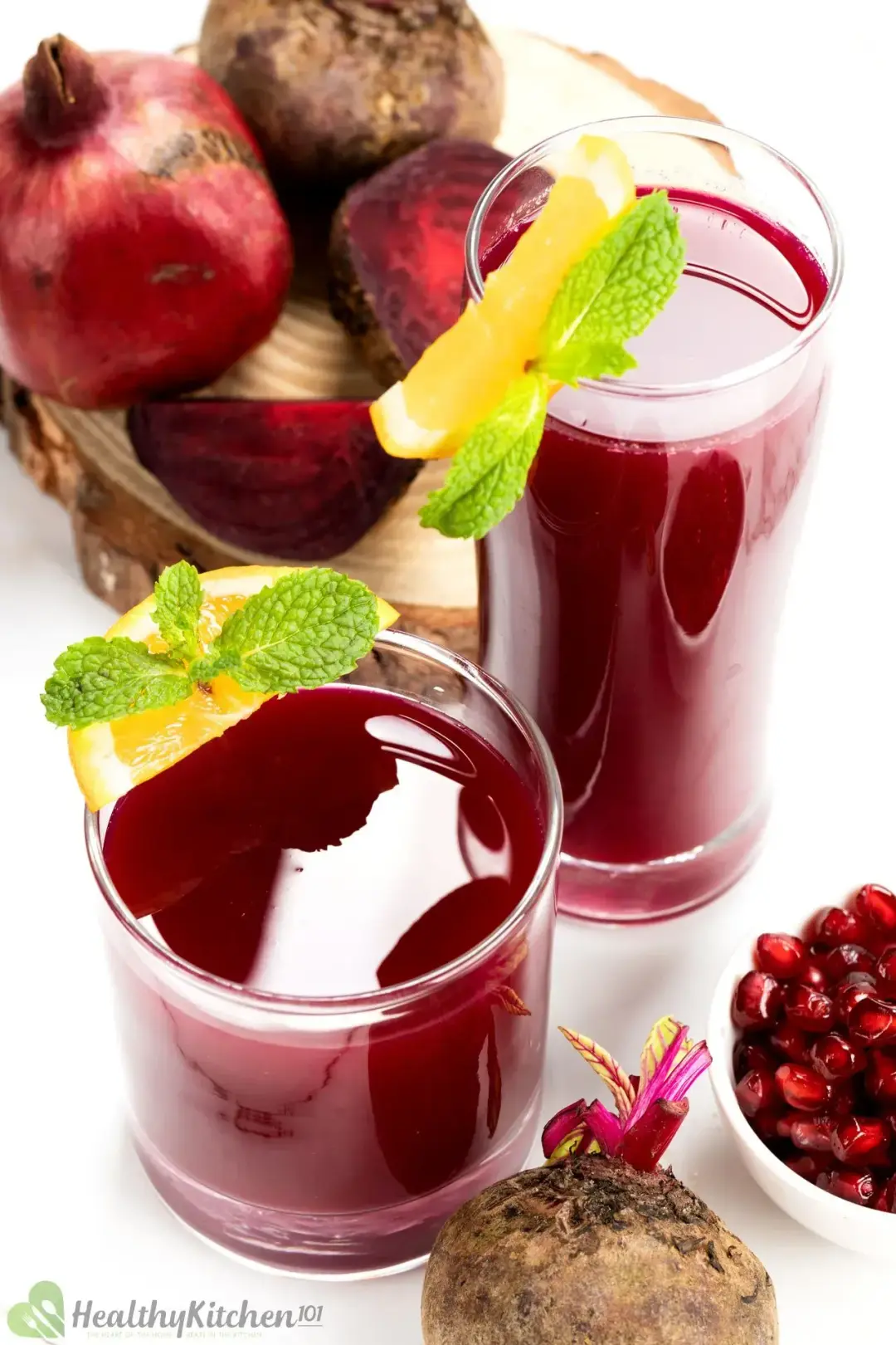 A short and a tall glass of beetroot juice, garnished with orange wedges and mints, next to pomegranates and beetroots