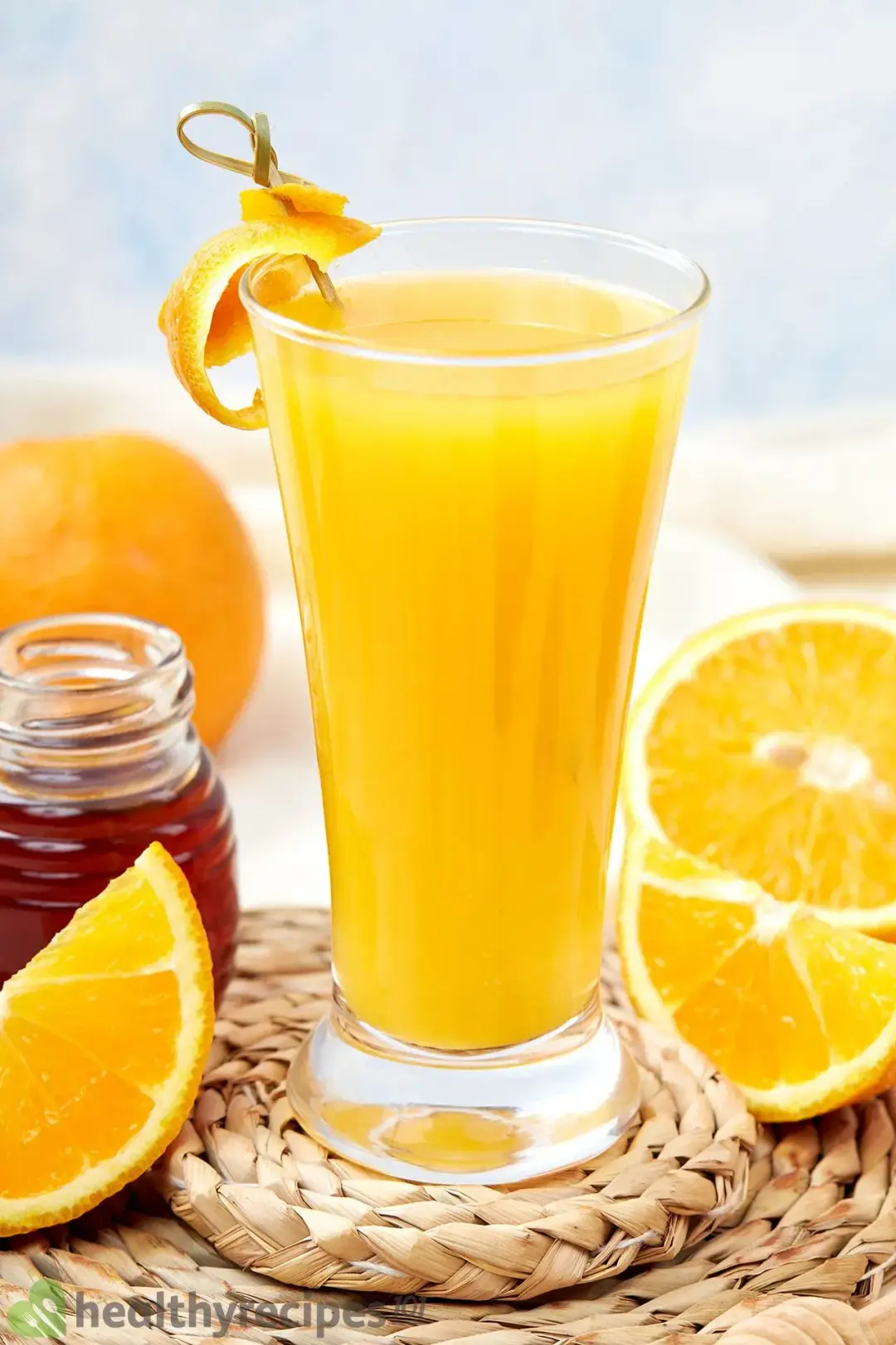 A tall glass filled with orange juice, next to some orange wedges and a jar of honey