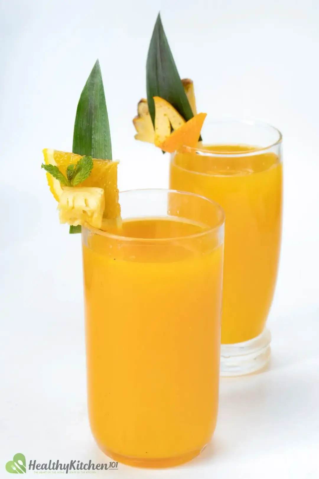 Two glasses of orange pineapple drinks garnished with a piece of pineapple leaf, orange, and pineapple