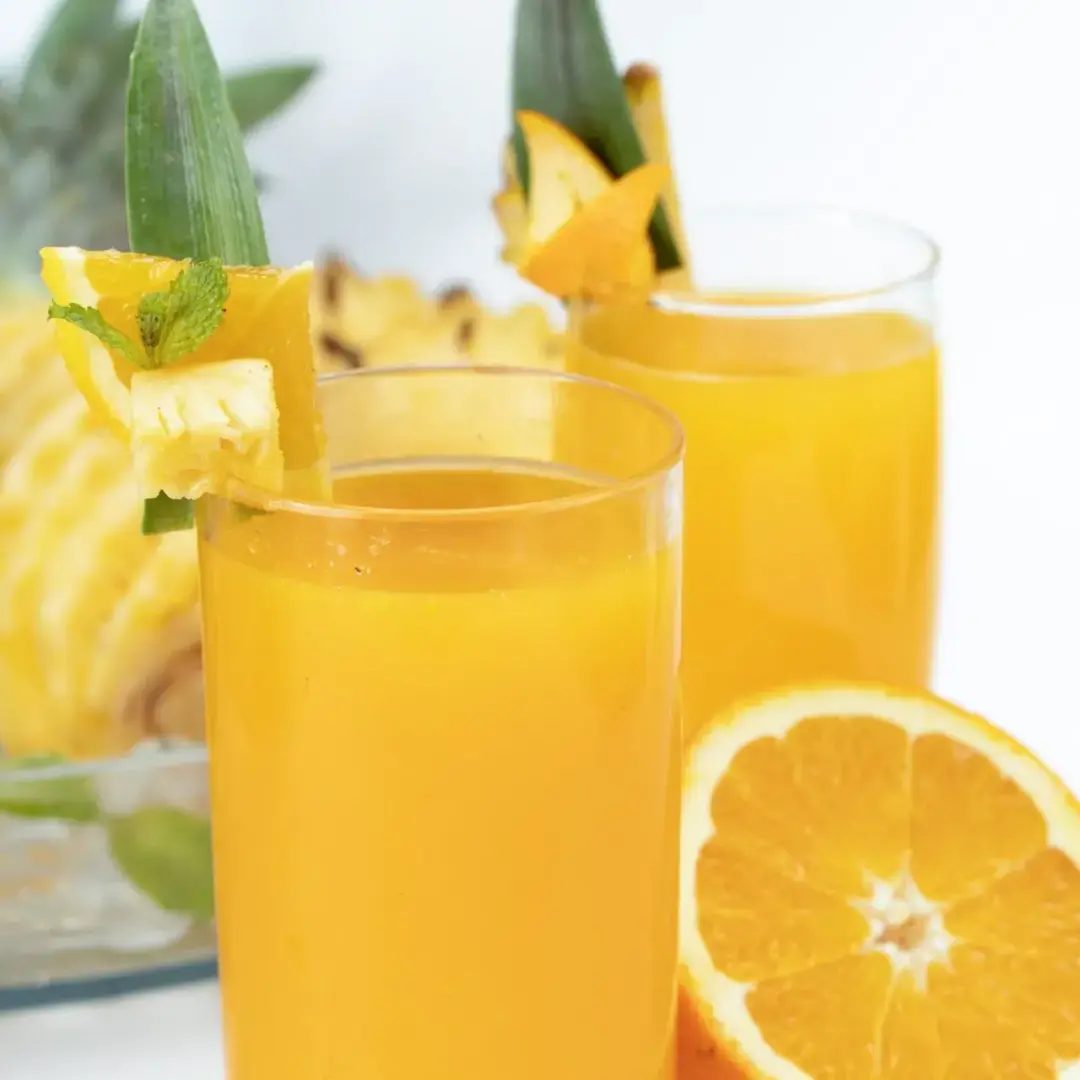 Two glasses of orange pineapple juice, next to half an orange and some peeled pineapples