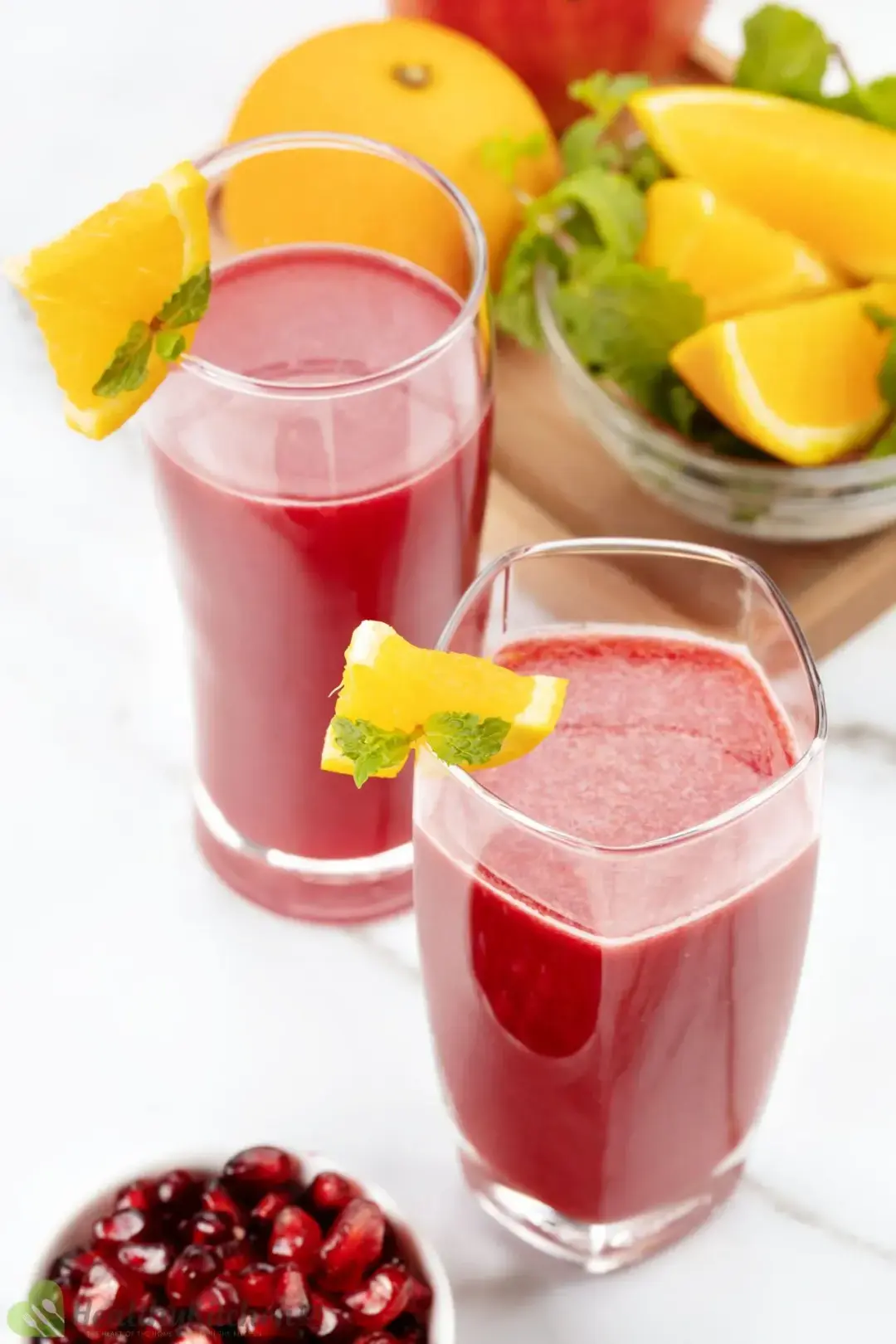 Two glasses of pomegranate orange juice put next to each other and a small bowl of pomegranate seeds