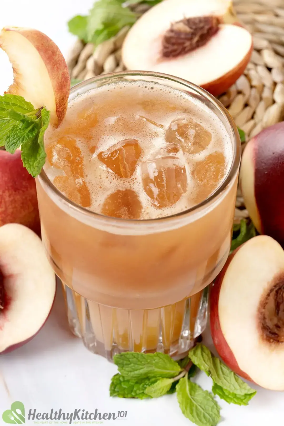 An iced glass of brown peach juice, next to peach halves and mint leaves