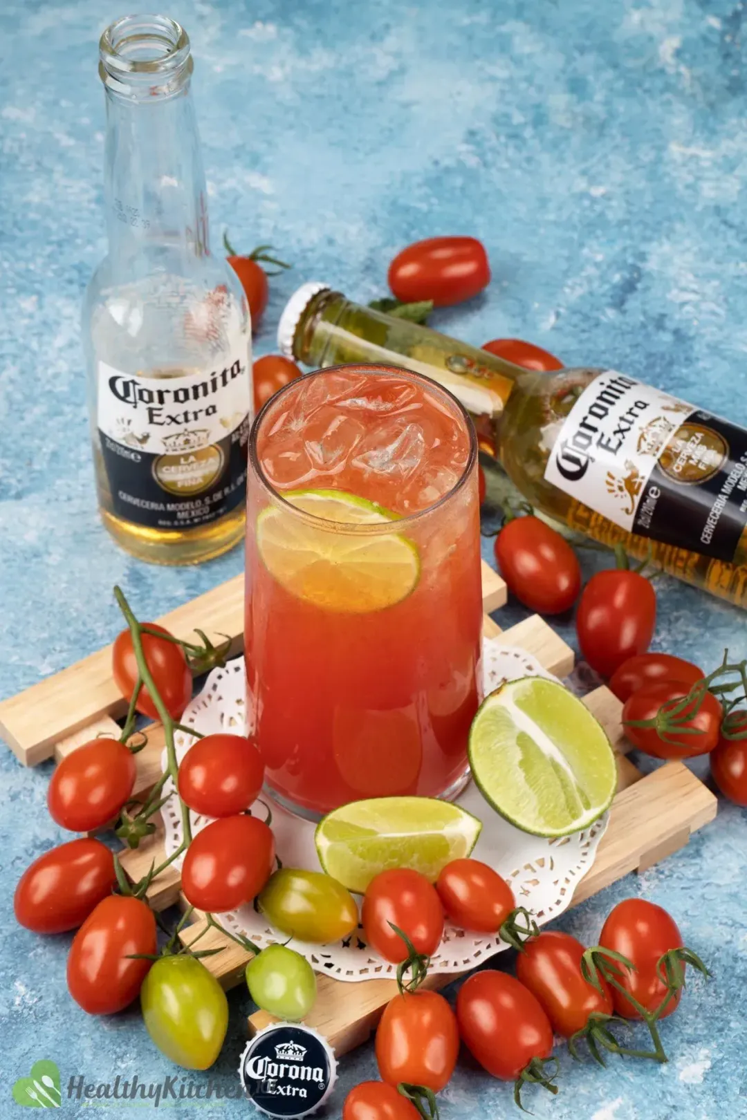 A glass of beer tomato cocktail surrounded by cherry tomatoes, halved limes, and beer bottles