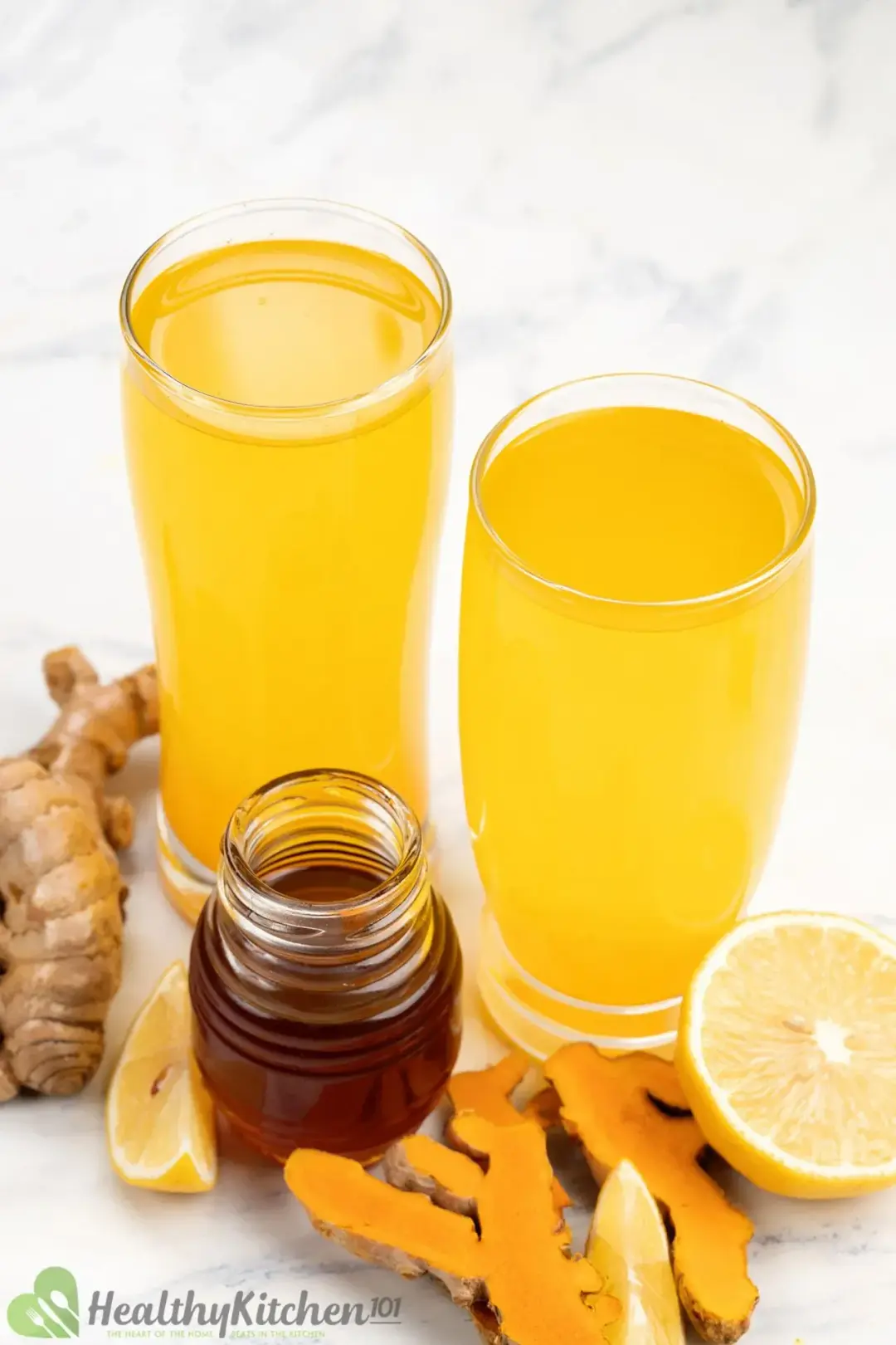 two glasses of turmeric and apple cider vinegar surrounded by turmeric roots, honey jar and half lemon