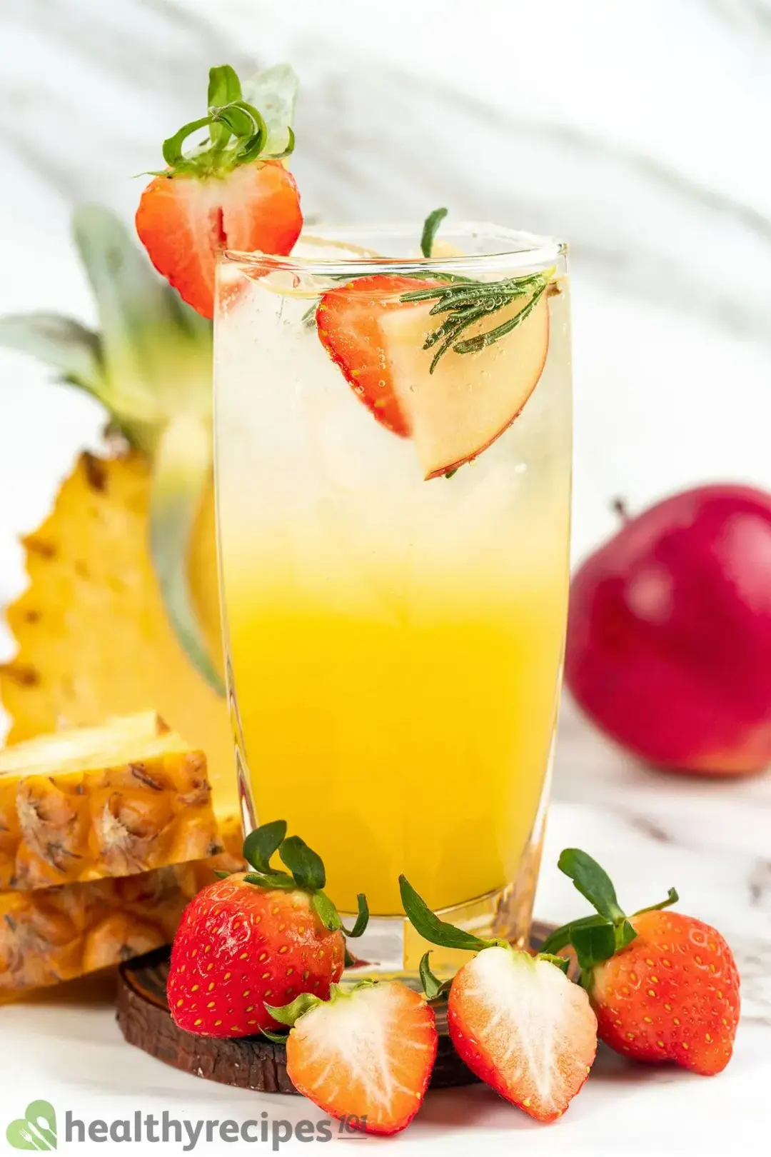 A tall glass of orange jungle juice cocktail with strawberries inside and around, next to pineapples and apples