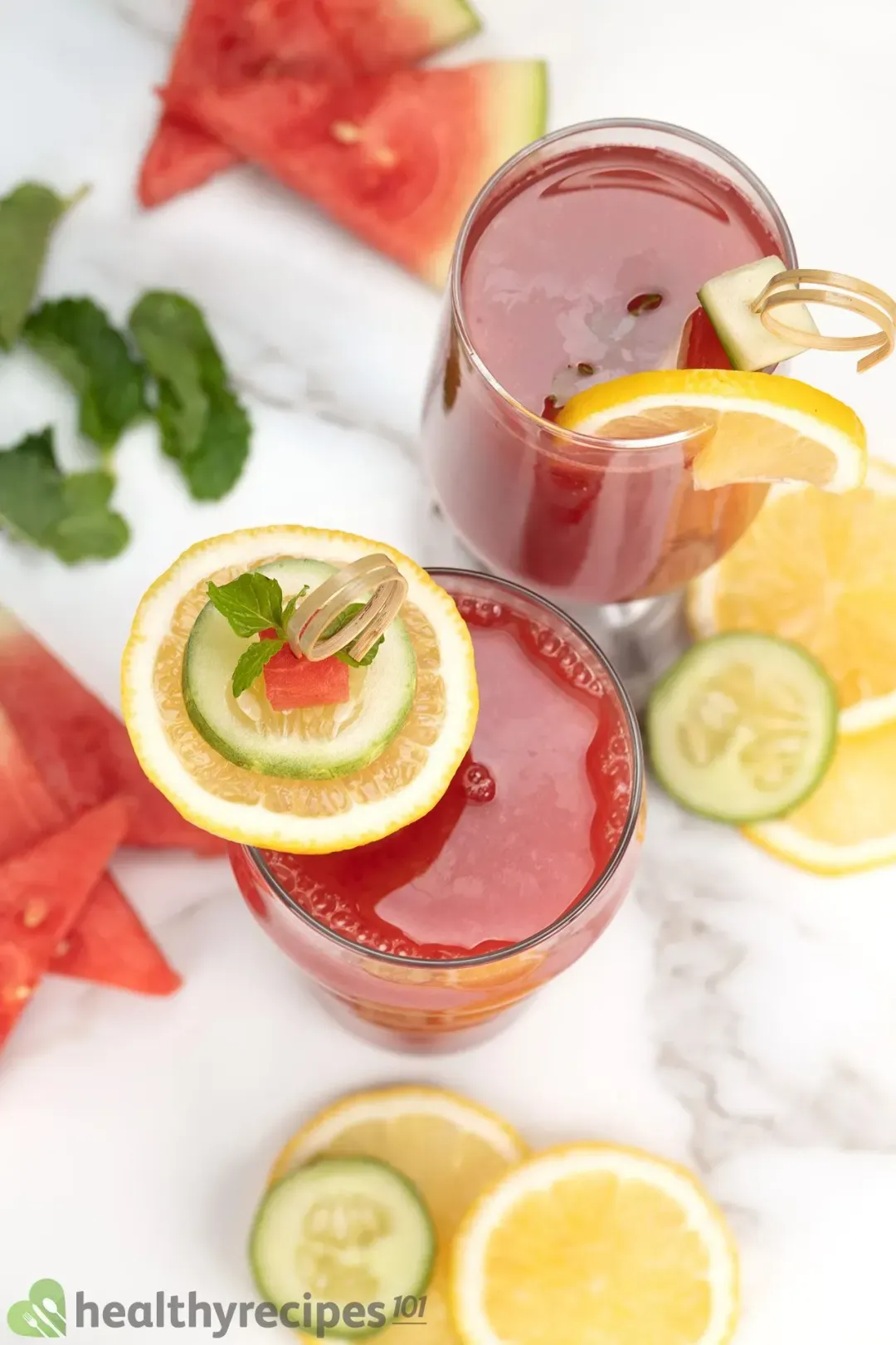 A high-angle shot of two red glasses of watermelon cucumber juice surrounded by lemon slices, cucumber slices, and watermelon slices