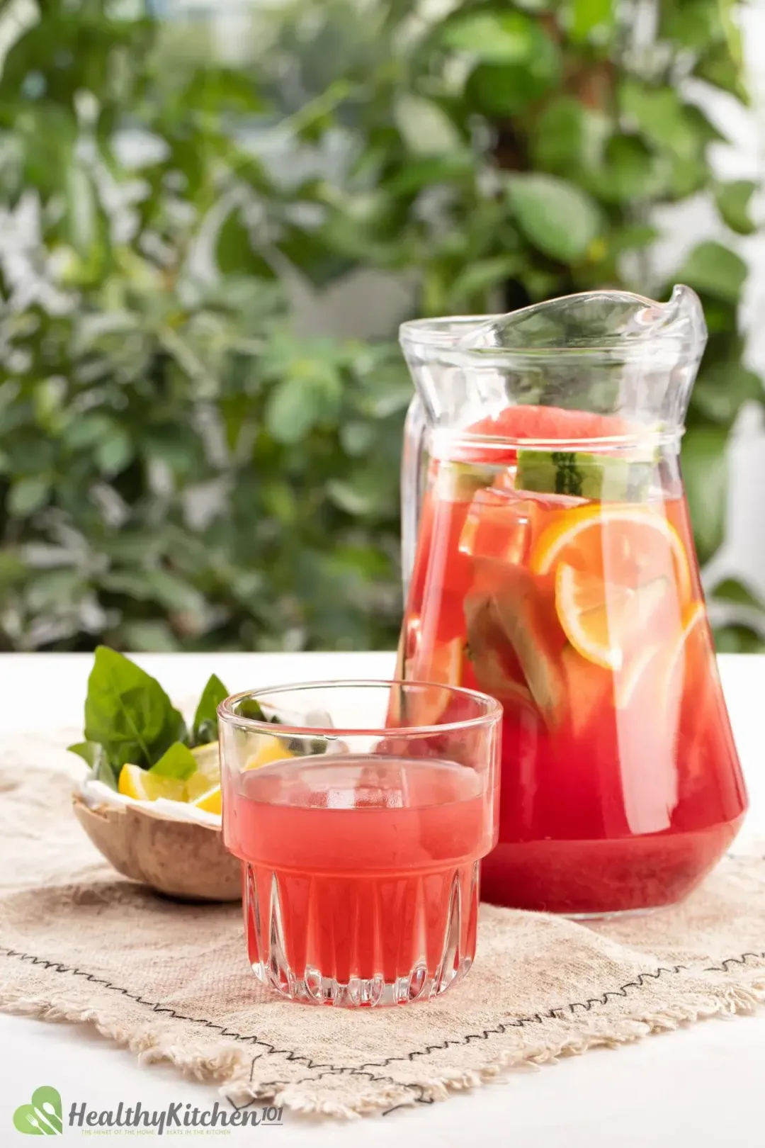 A picture of a jug and a glass of watermelon jungle juice placed on a rug next to a coconut shell with lemon slices and basil leaves in it