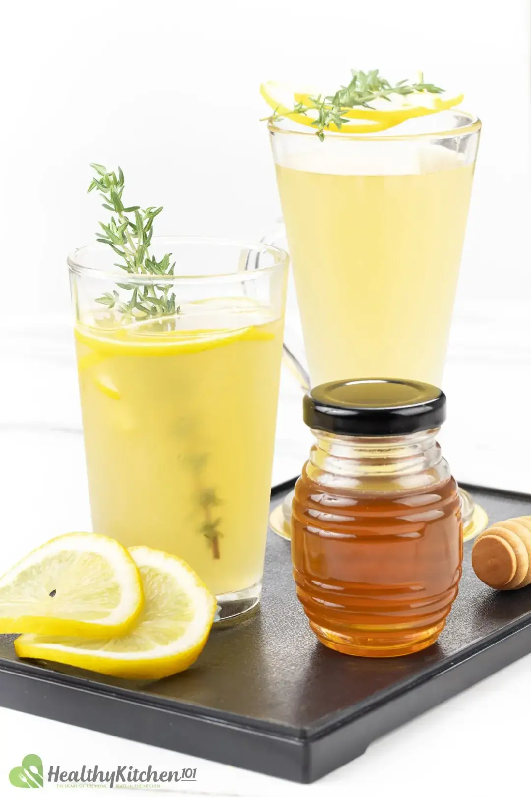 Two glasses of lemonade with thyme sprigs next to lemon wheels and a honey jar