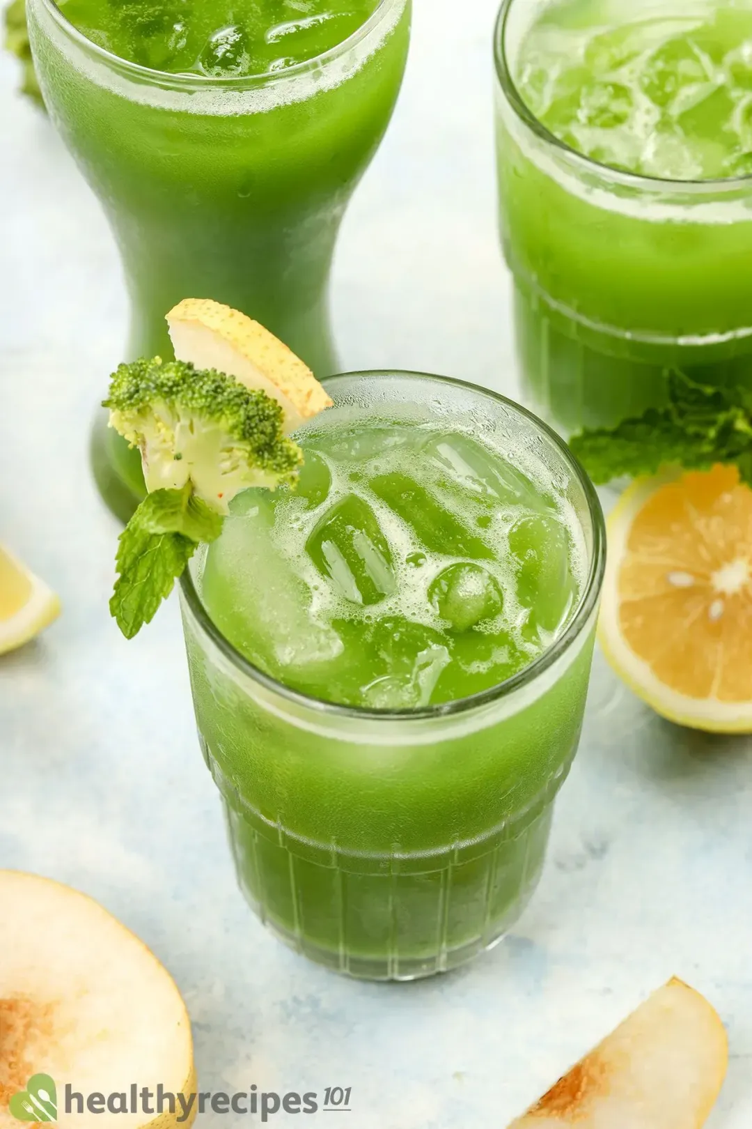 Three iced glasses of broccoli juice drinks garnished with lime wedges, mints, and next to some fruits
