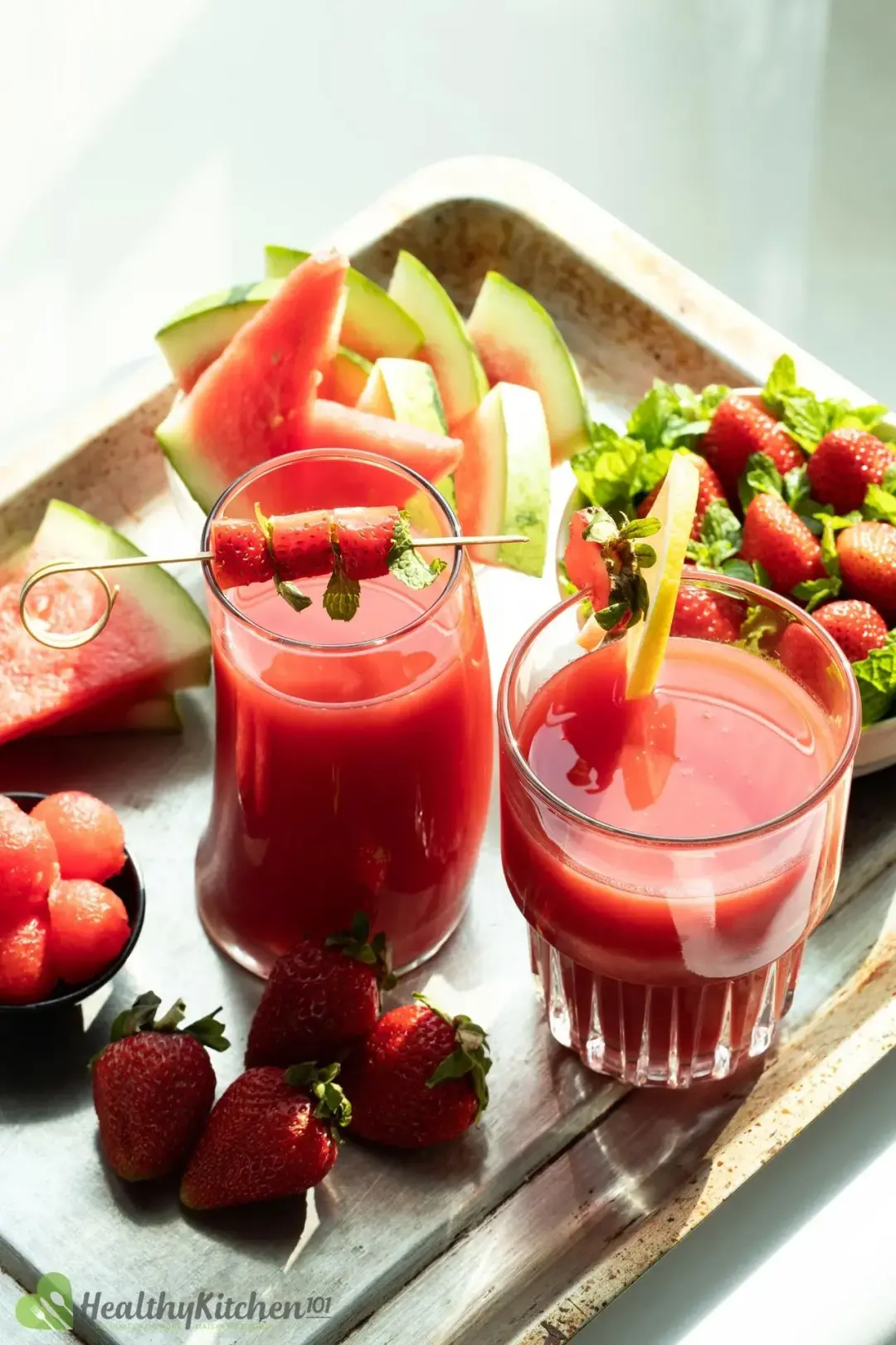 Two glasses of red juice in a steel tray decorated with strawberries and lemon surrounded by strawberries and watermelon