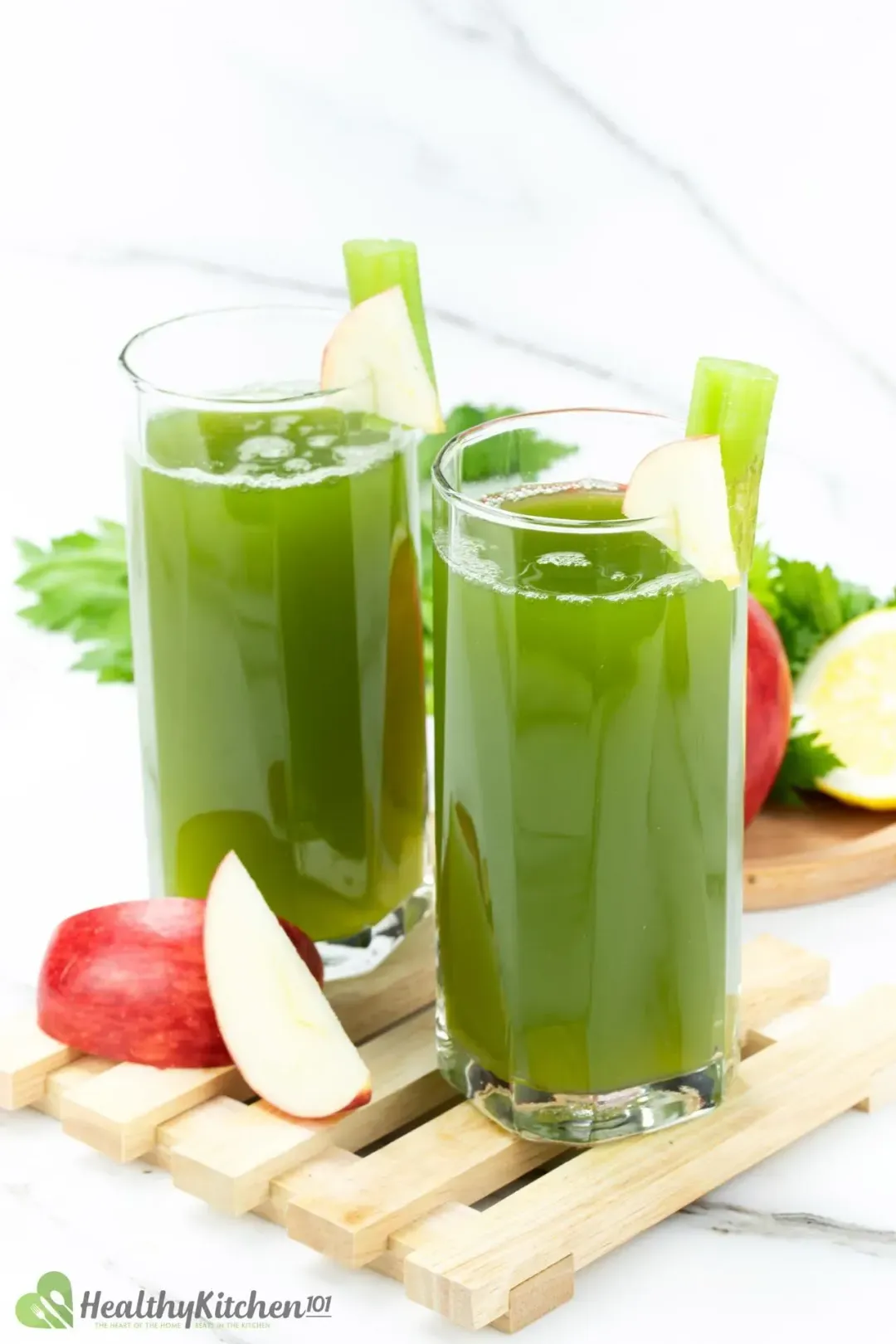 Two glasses of celery juice put on wooden bars, next to red apple wedges and greens
