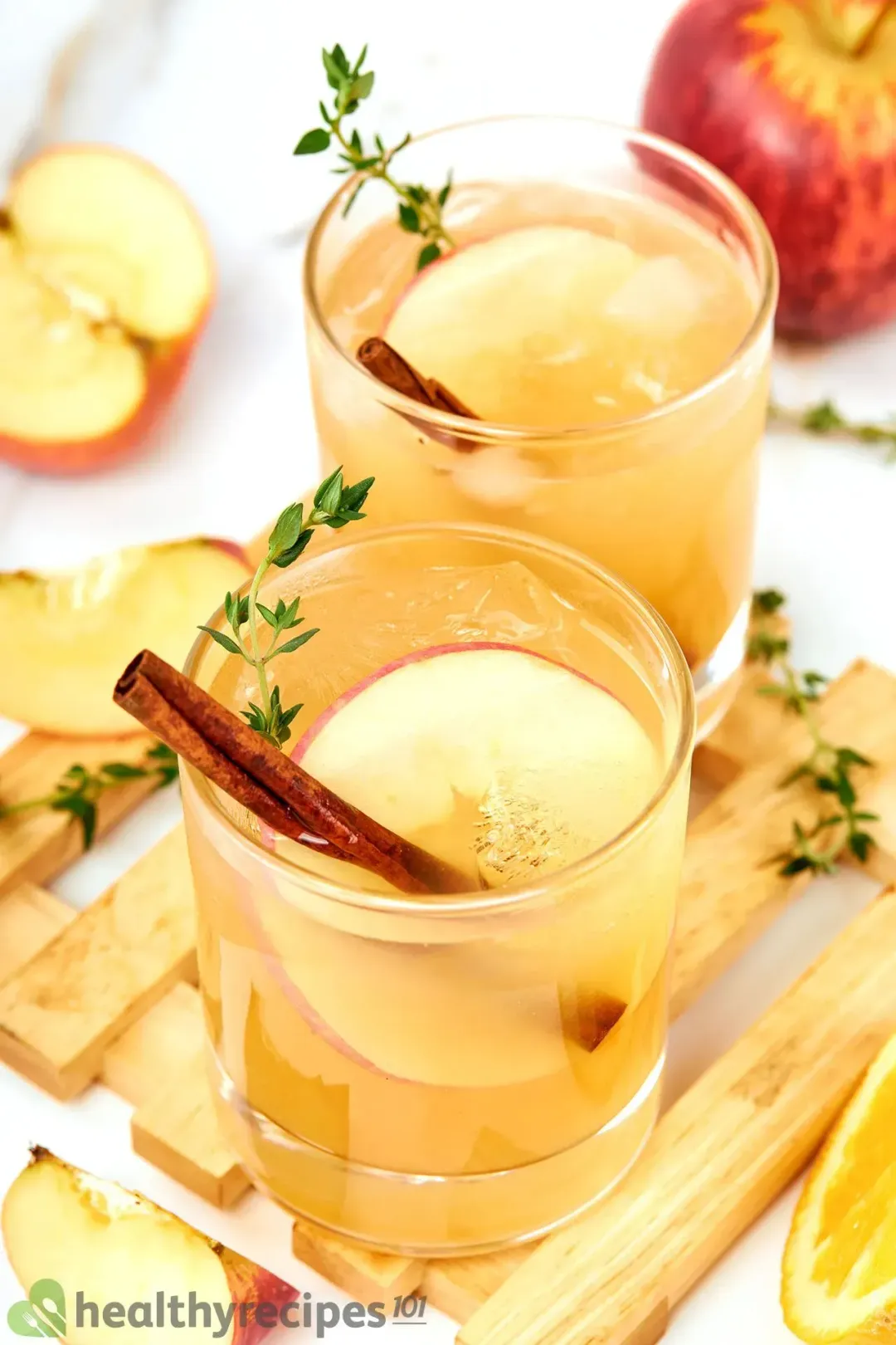 Two short glasses of apple cider cocktails and cinnamon sticks, apple slices, and rosemary, next to some apple wedges