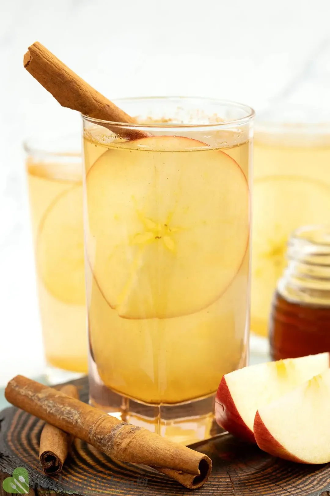 A shot glass of apple vinegar honey with cinnamon sticks, red apple wedges, and a honey jar